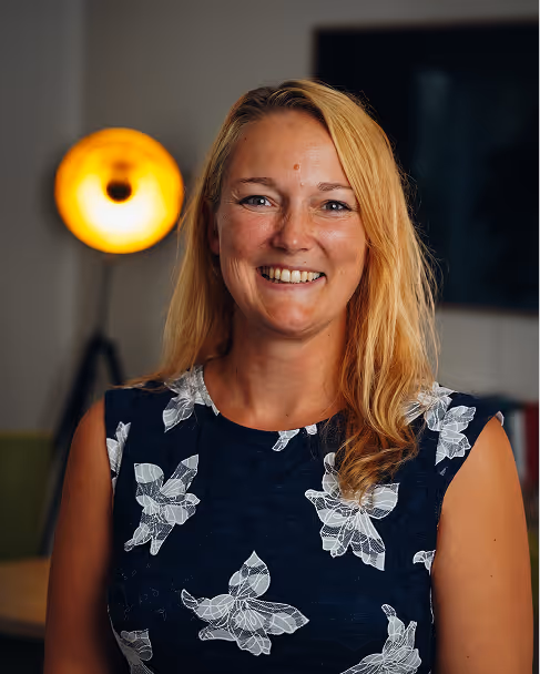 Smiling woman with blonde hair wearing a black sleeveless top with white floral patterns, standing indoors with a warm light in the background.