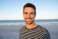 Smiling man with dark hair and a beard wearing a black and white striped shirt standing outdoors near the beach with the ocean and clear sky in the background.