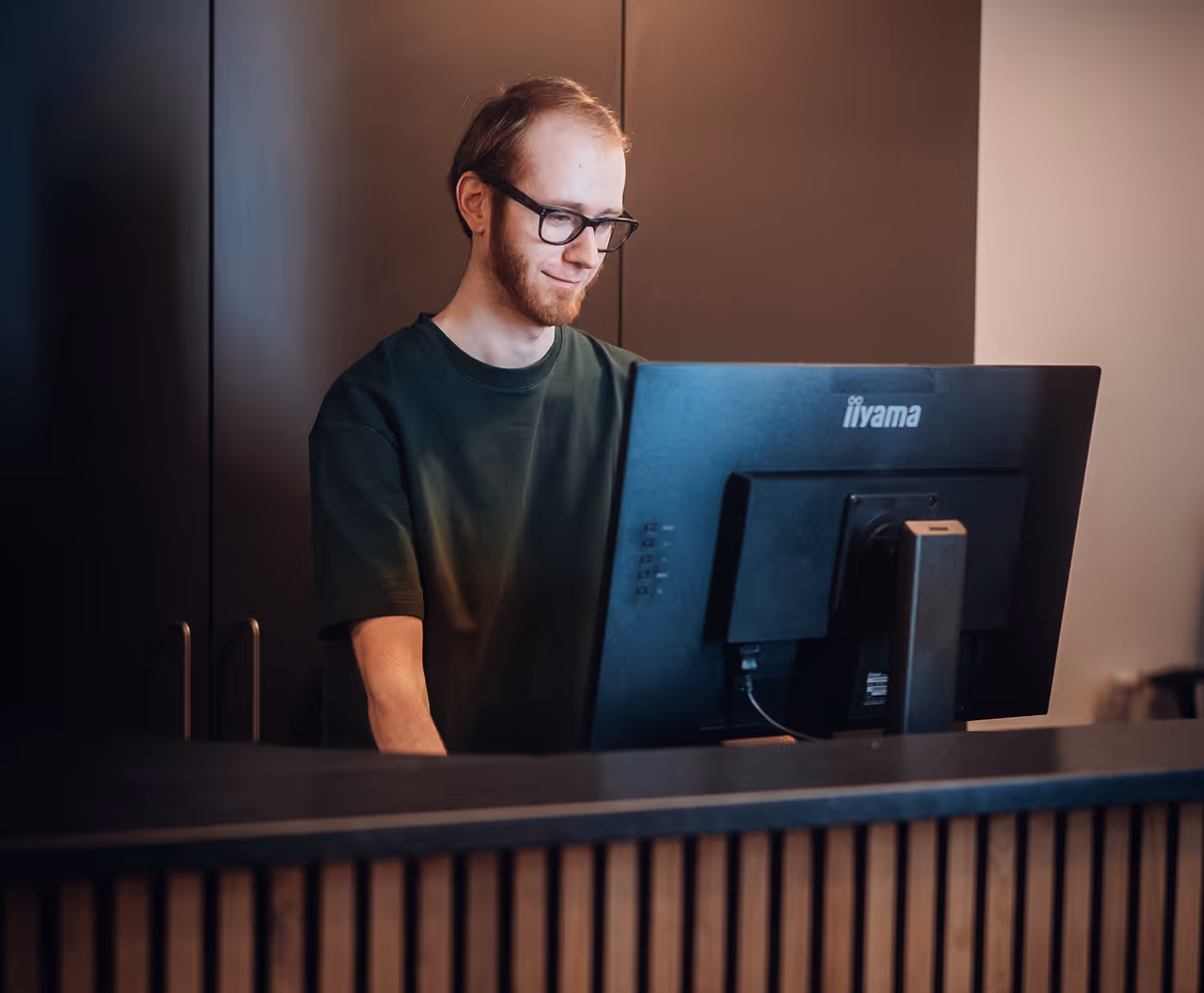 Young man with glasses smiling while working on a desktop computer behind a wooden counter.