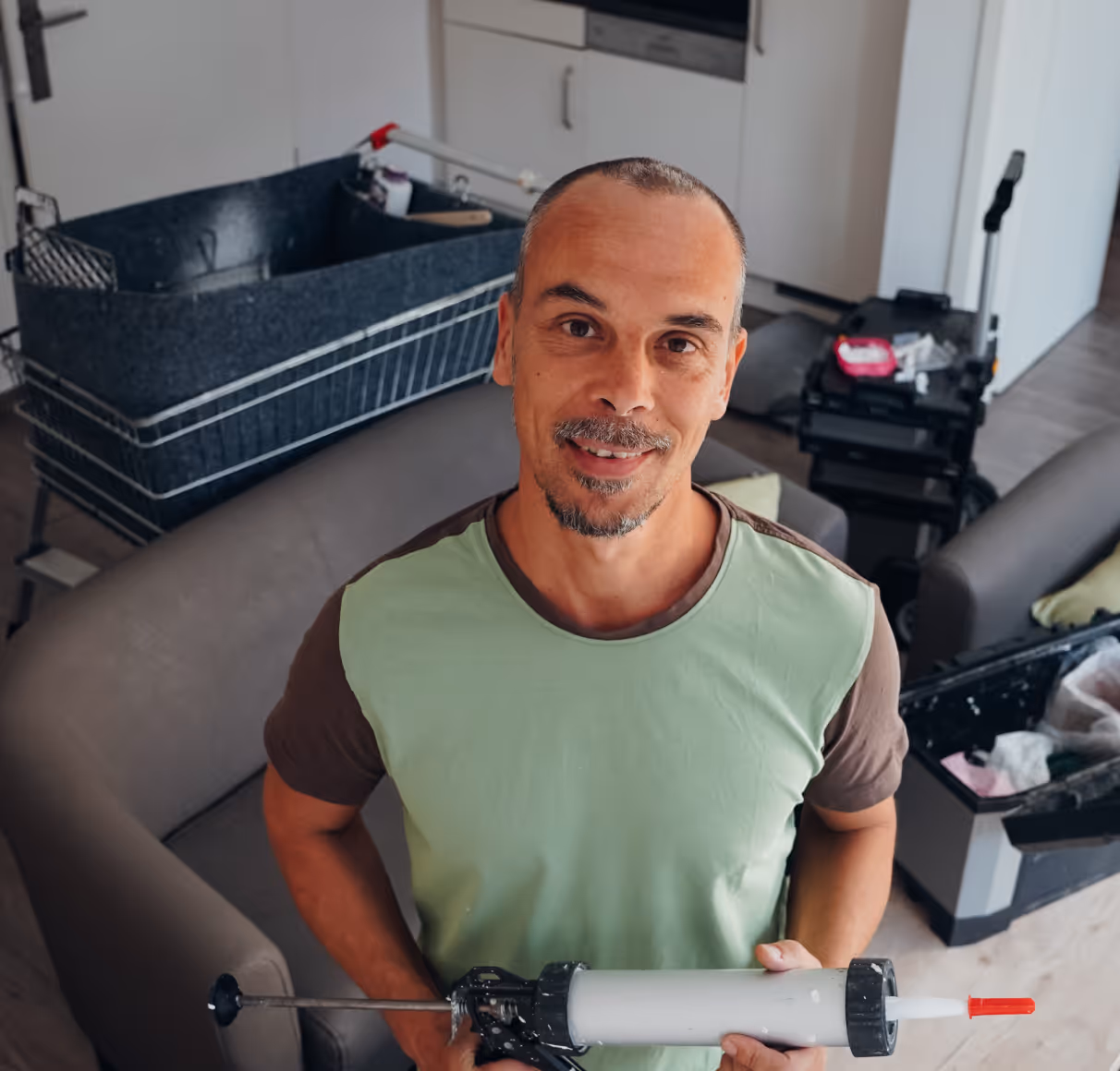 Smiling man with short hair holding a caulking gun in a living room with furniture and tools around.