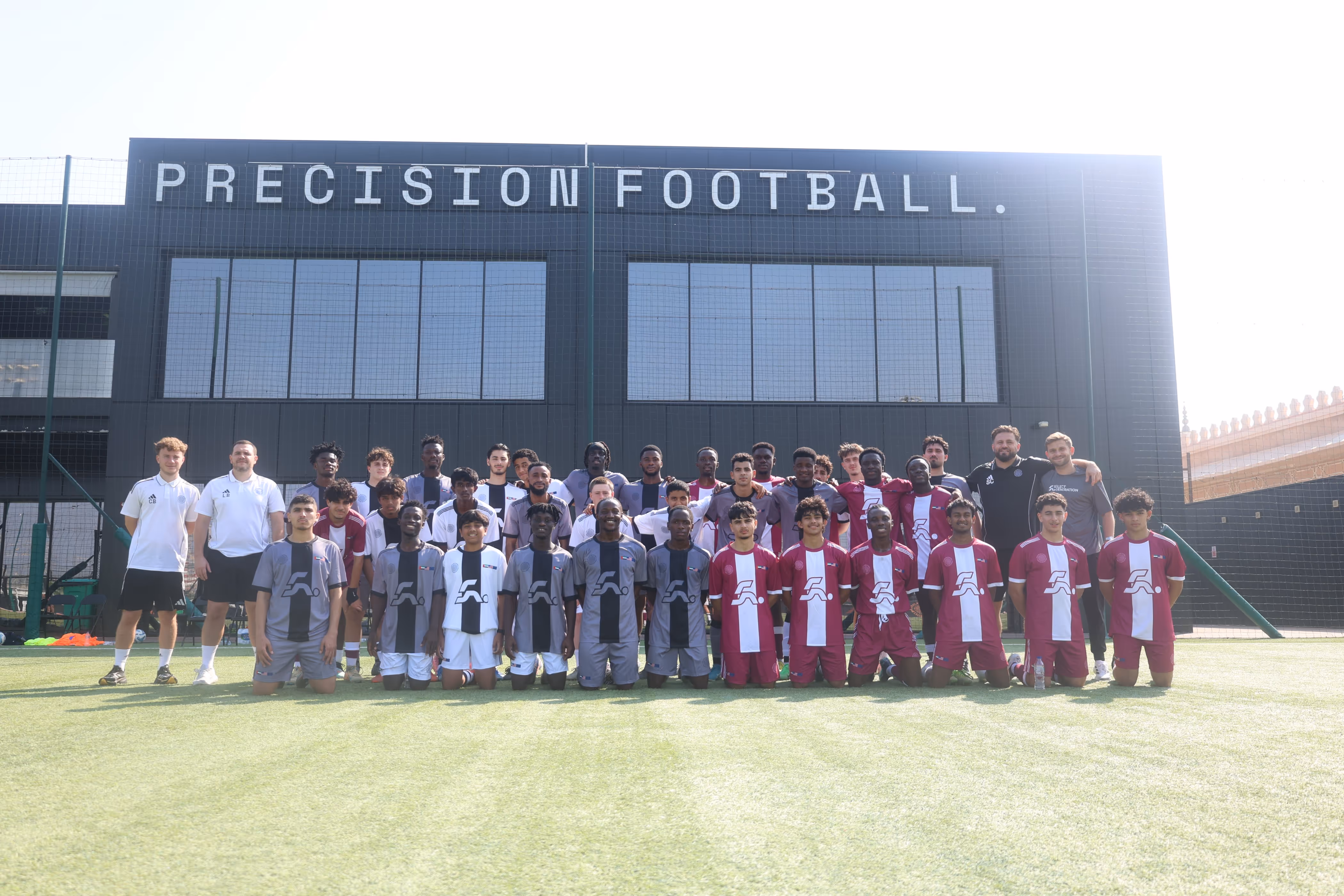 Group of young male soccer players and coaches posing on a field in front of a building with the sign 'PRECISION FOOTBALL.'