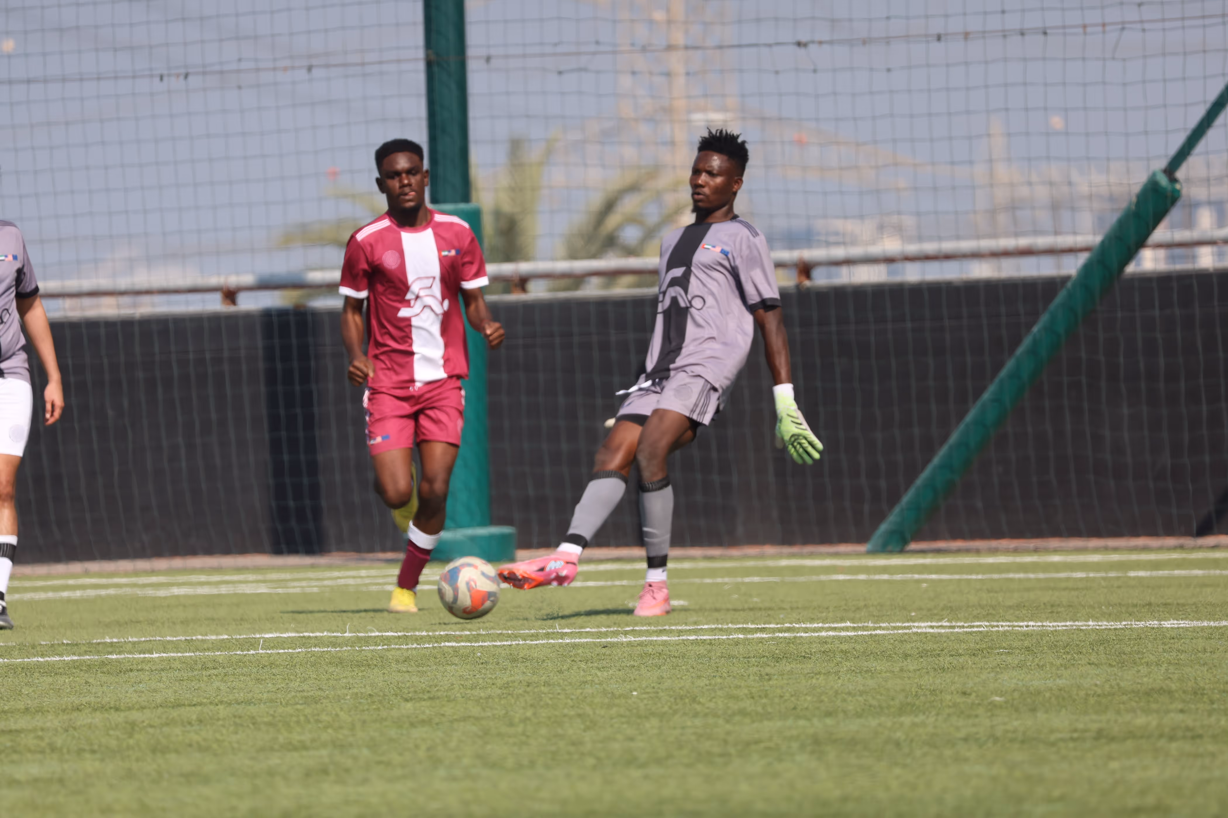 Two soccer players, one in a maroon and white uniform and the other in gray with green gloves, contesting near the ball on a green field.