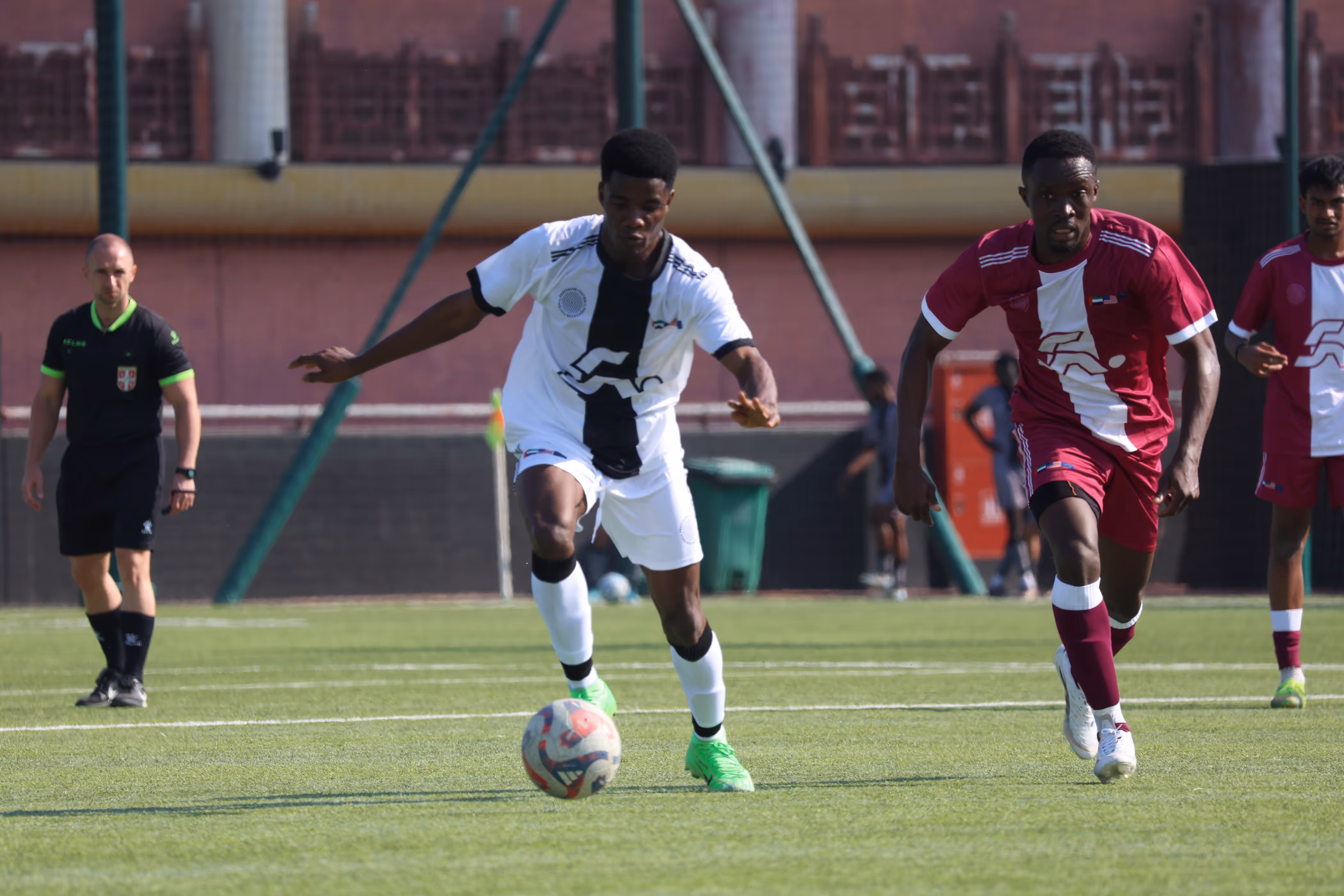 Two soccer players in white and maroon uniforms running towards a soccer ball on a grassy field during a match, with a referee in the background.
