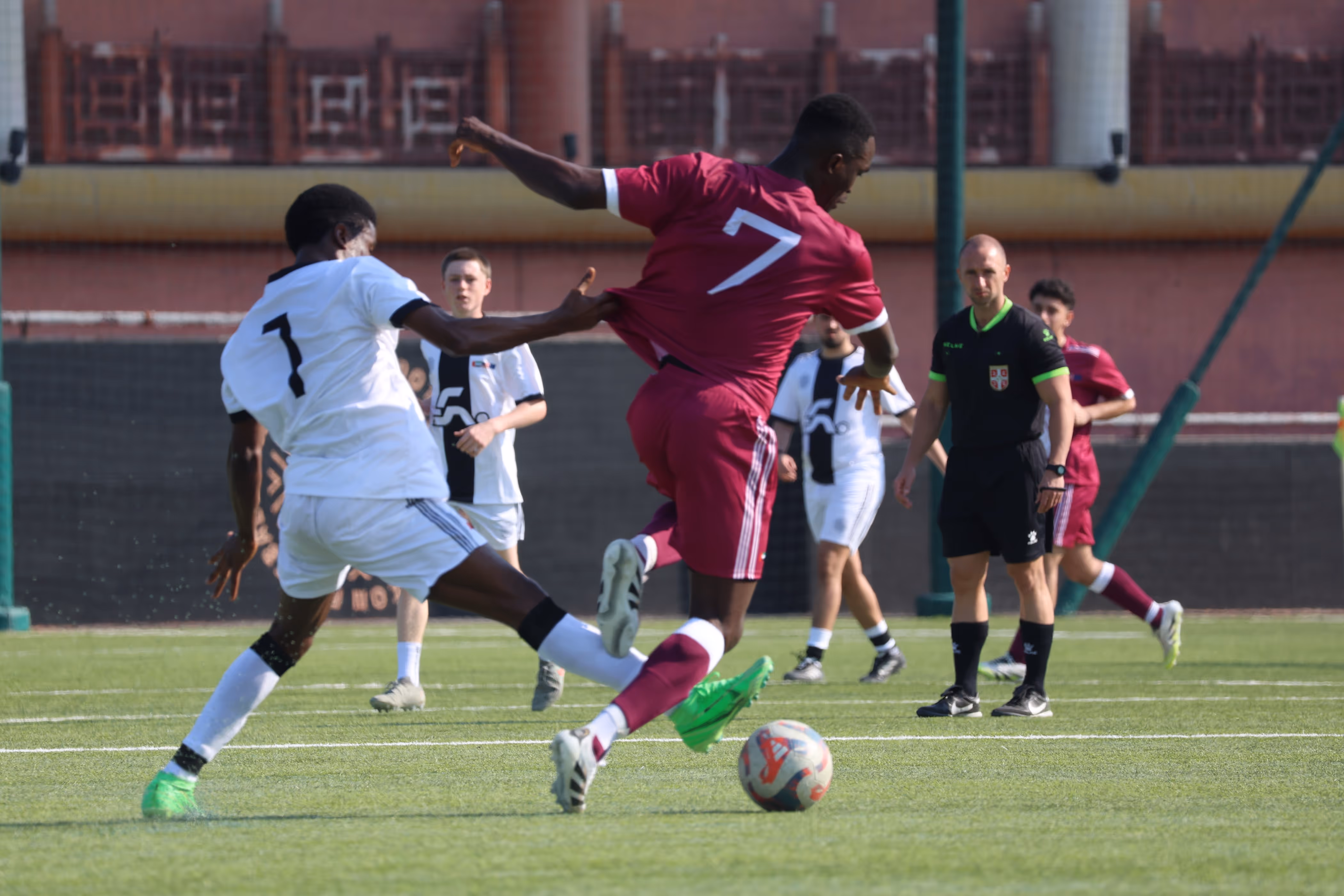 Soccer players from two teams jumping to head the ball on a green field, with players in maroon and white uniforms.