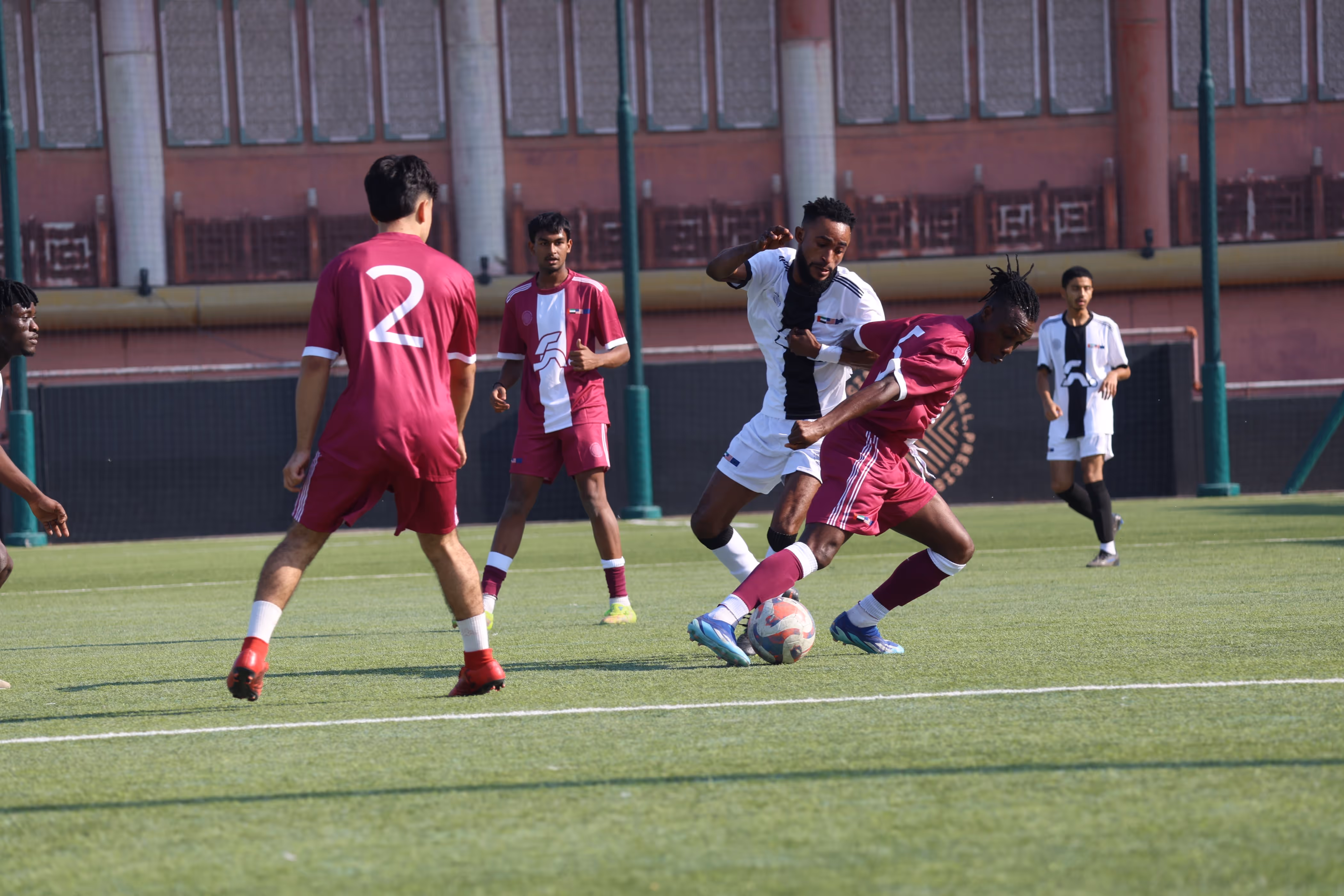 Soccer players from two teams jumping to head the ball on a green field, with players in maroon and white uniforms.