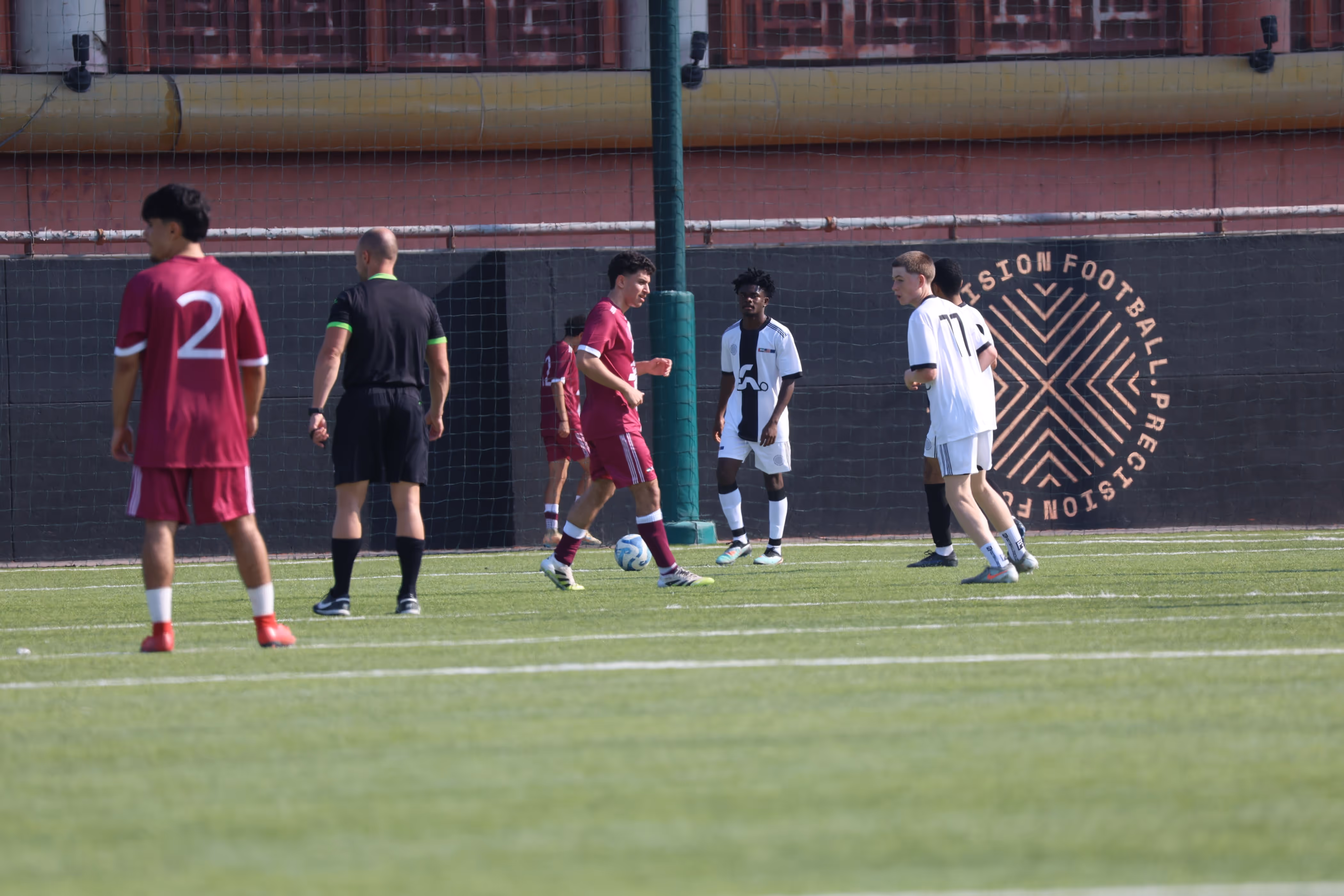 Soccer players from two teams jumping to head the ball on a green field, with players in maroon and white uniforms.