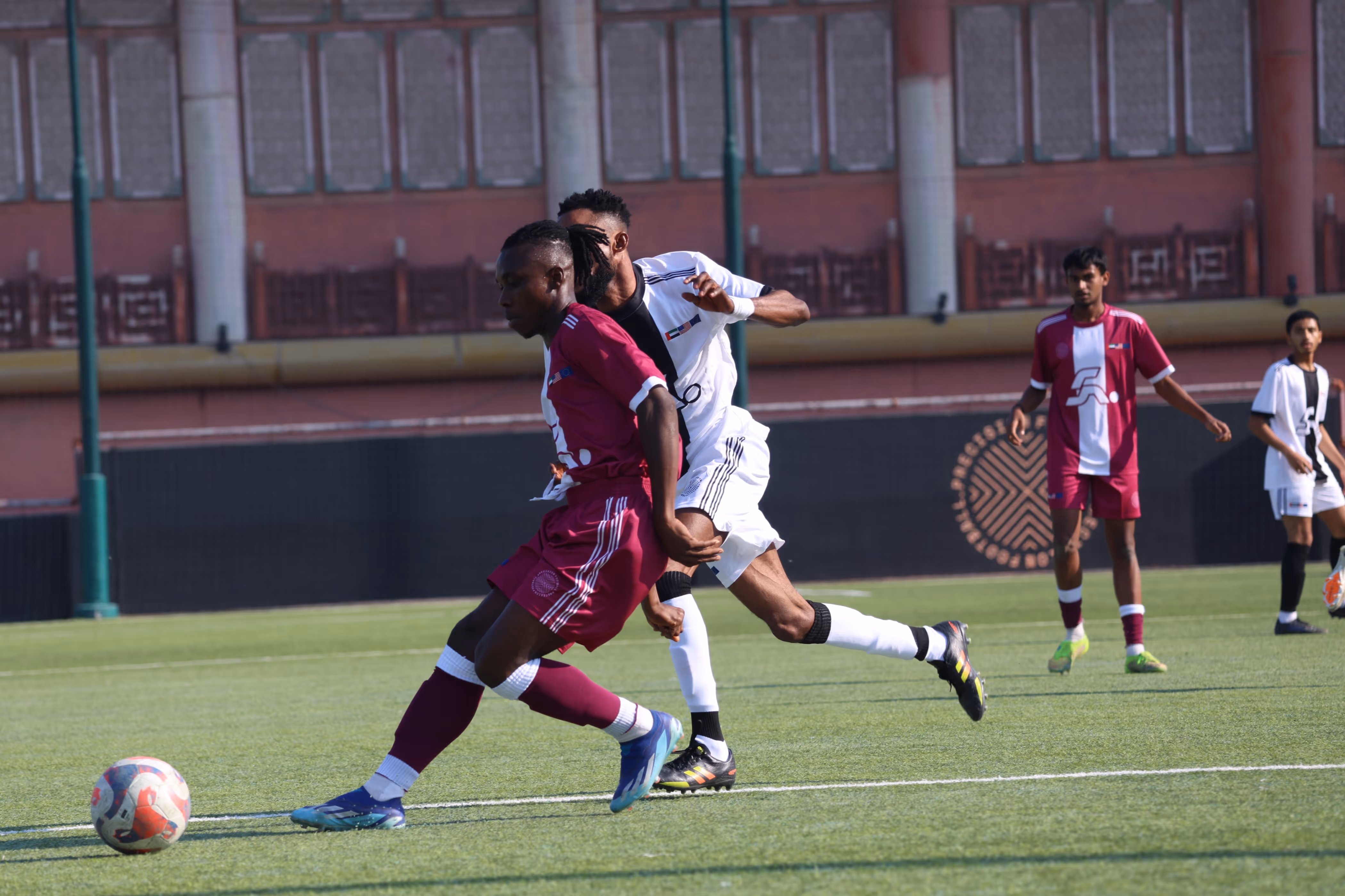Two soccer players in maroon and white jerseys contest a ball on a green field with two additional players in the background.