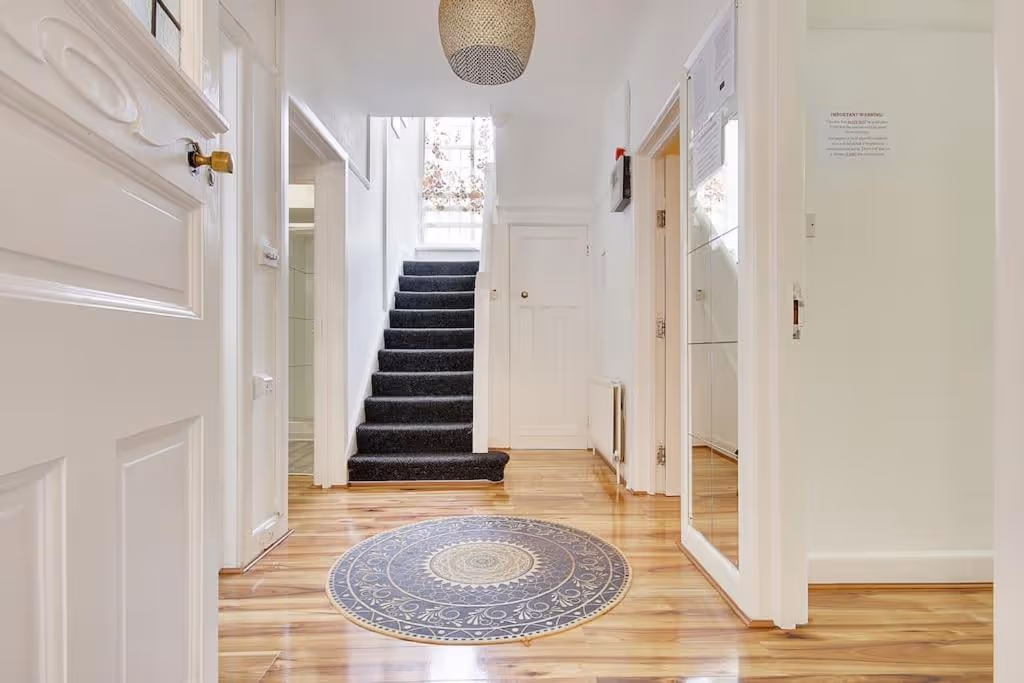 Bright hallway with wooden floor, a round decorative rug, white walls, and carpeted stairs leading upstairs.