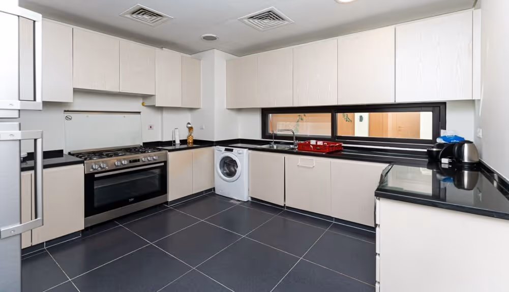 Modern kitchen with light wood cabinets, black countertops, stainless steel stove, washing machine, and a wide horizontal window above the sink.
