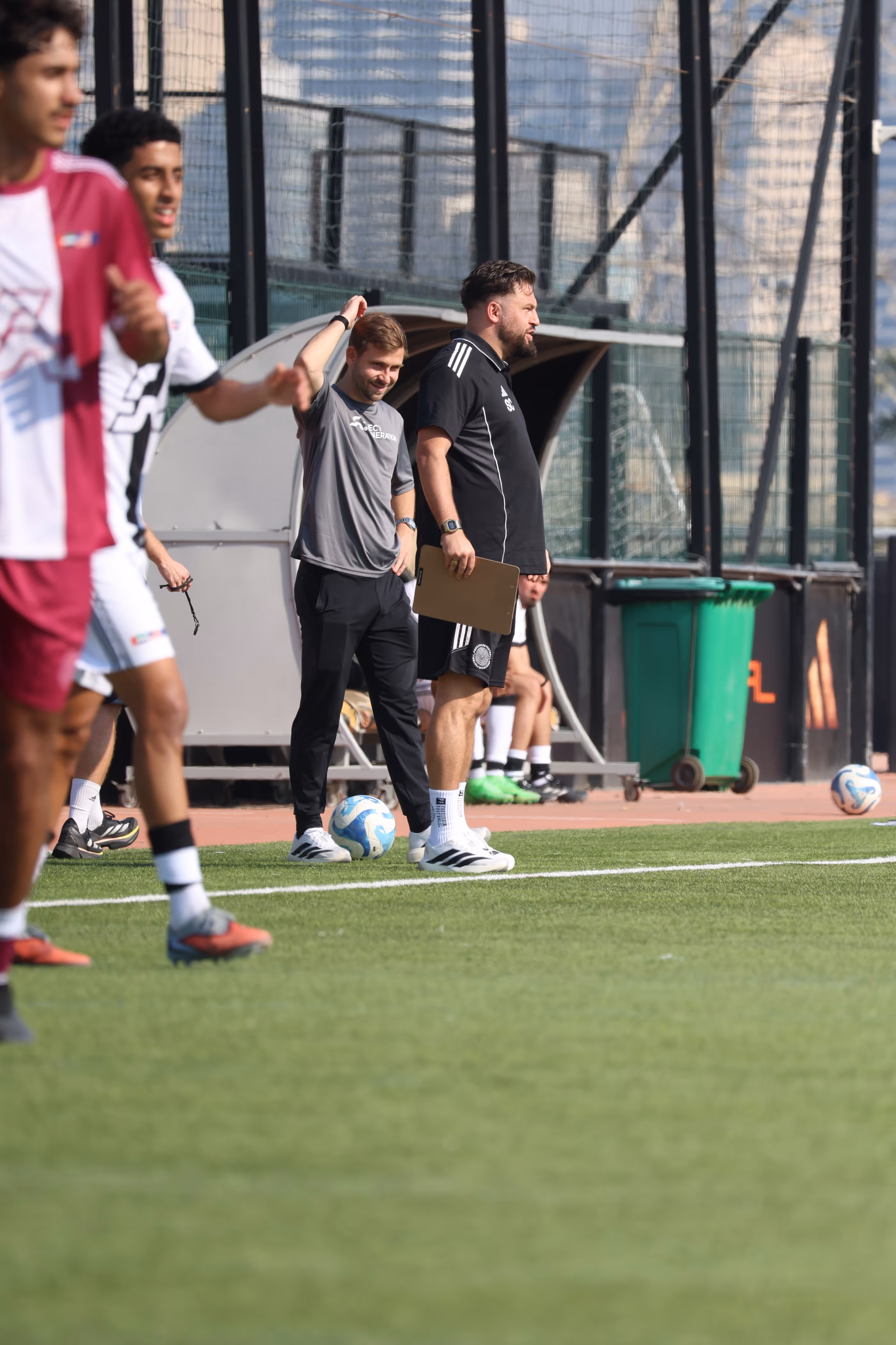 Two coaches stand on the sidelines of a soccer field, one holding a clipboard and the other with a soccer ball by his feet, with players partially visible in the foreground.