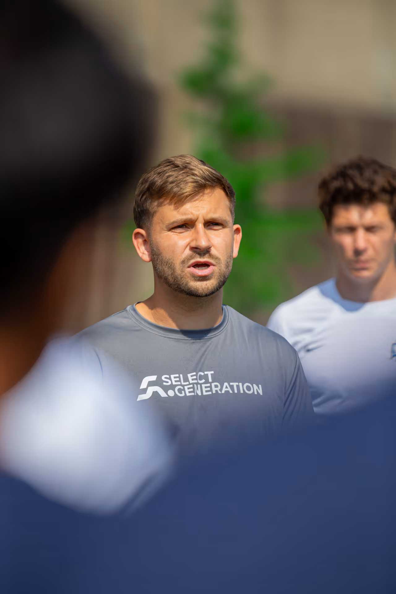 Man in a gray Select Generation t-shirt speaking outdoors with blurred people in foreground and another person in white shirt in background.