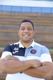 Smiling man with arms crossed wearing a black, white, and gray polo shirt standing outdoors with bleachers in the background.