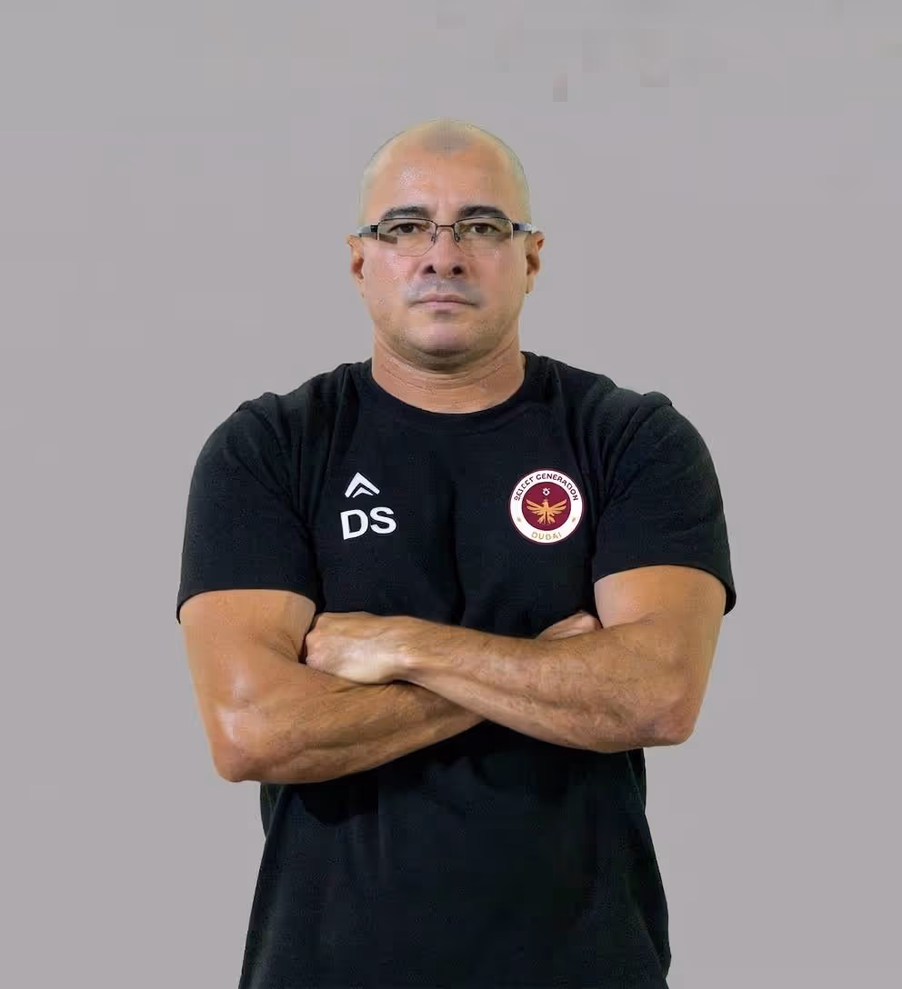 Man with arms crossed wearing a black Adidas t-shirt and a black cap against a plain light background.