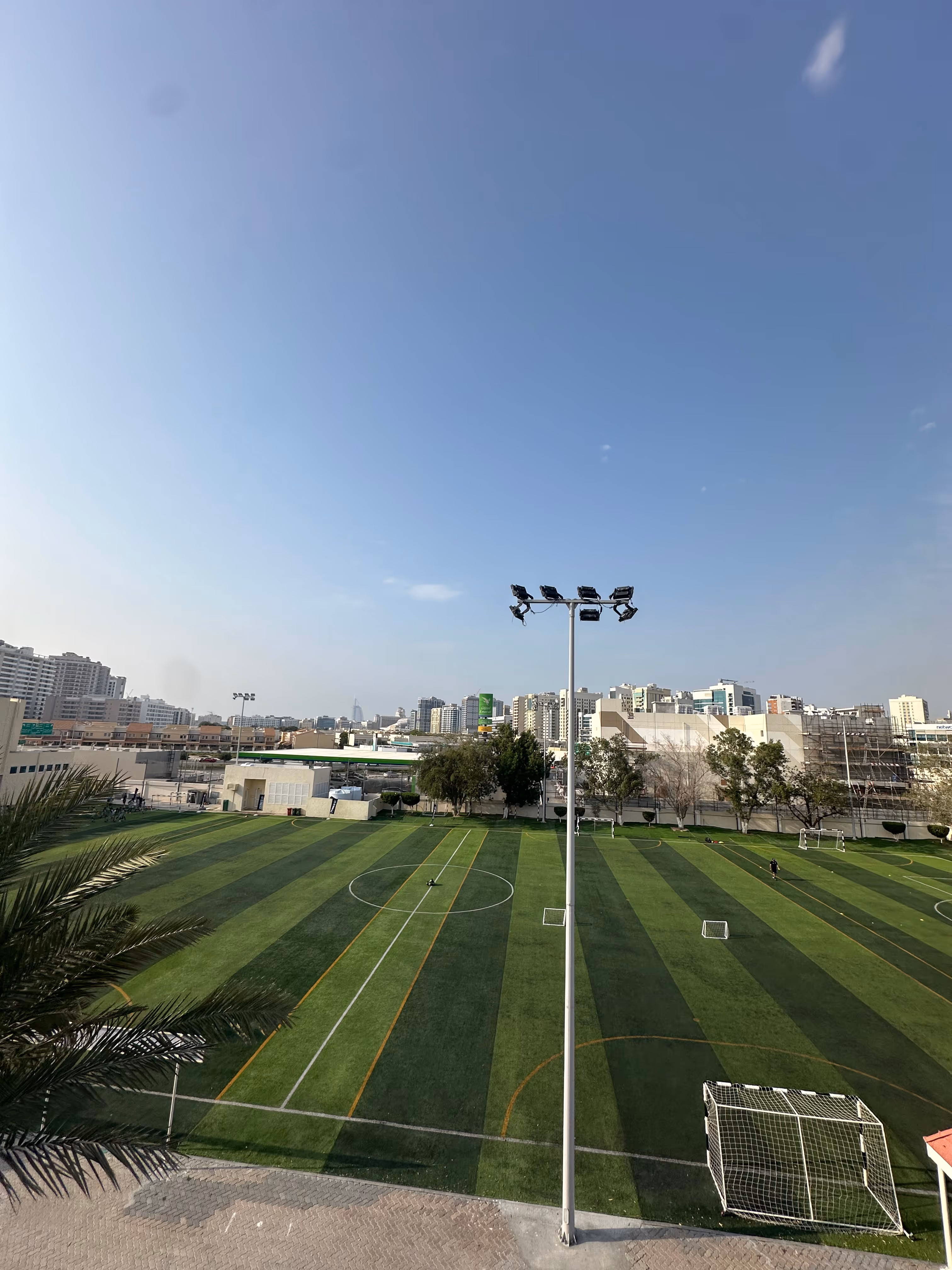 Outdoor sports field with green artificial turf, three soccer goals, a tall light pole, and surrounding buildings under a clear sky.