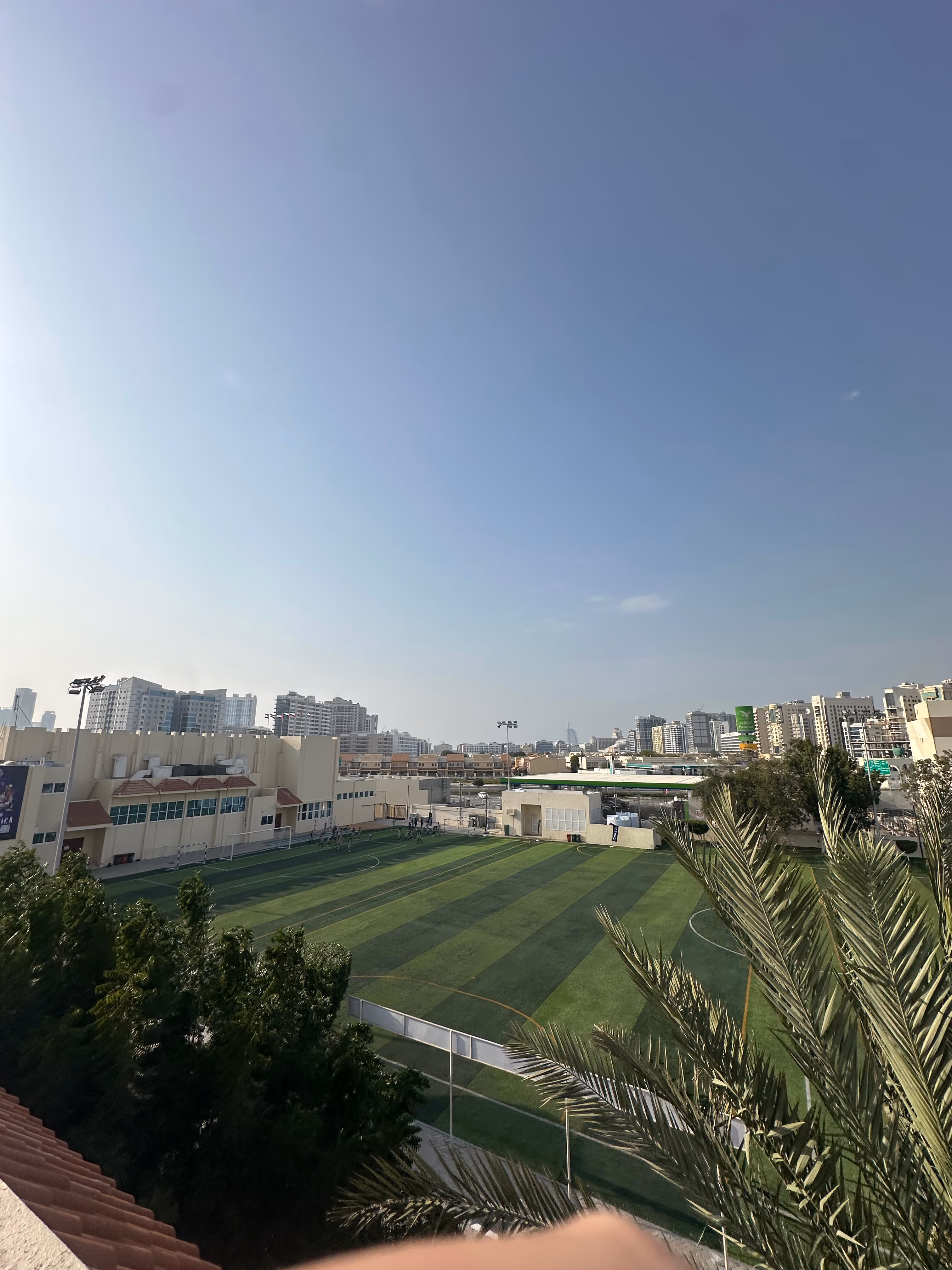 Outdoor sports field with green artificial turf, three soccer goals, a tall light pole, and surrounding buildings under a clear sky.