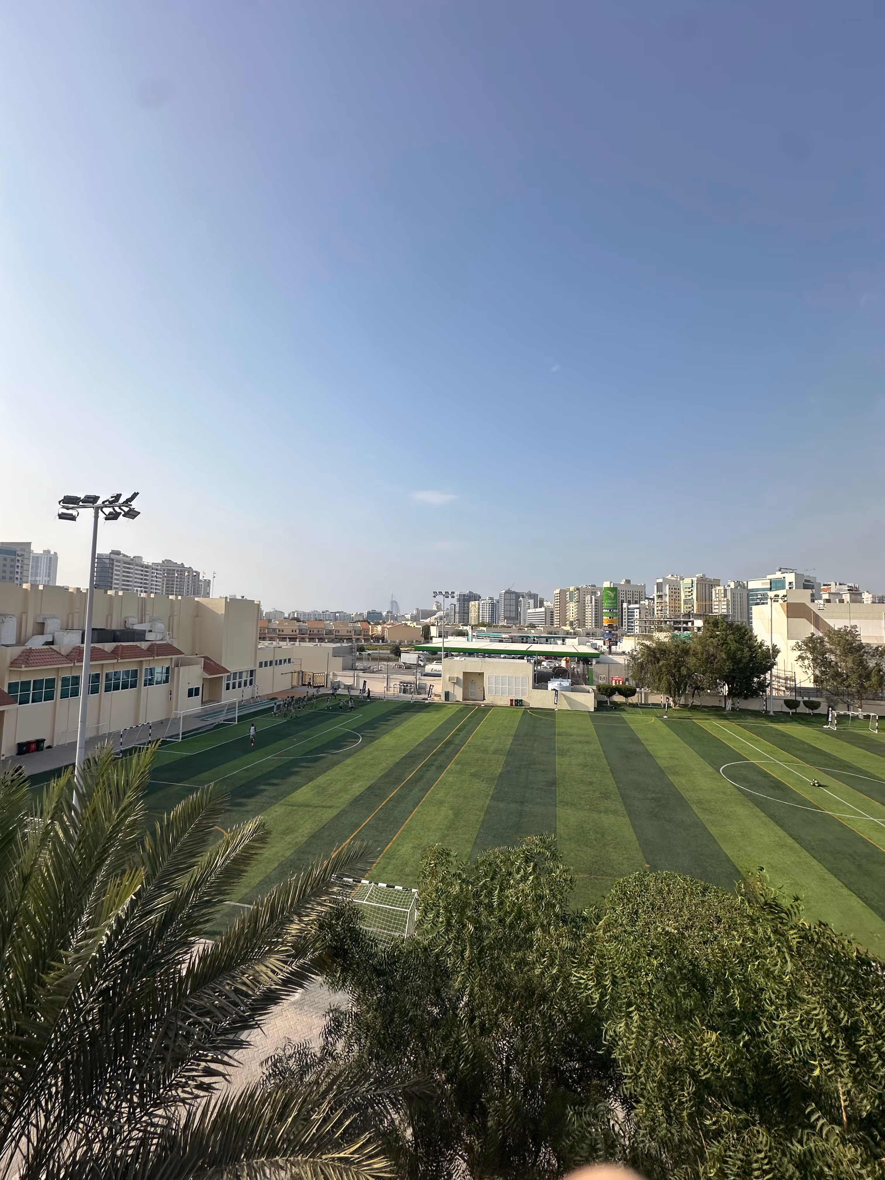 A large, green artificial turf soccer field with white and yellow lines, surrounded by buildings and trees, under a clear blue sky.