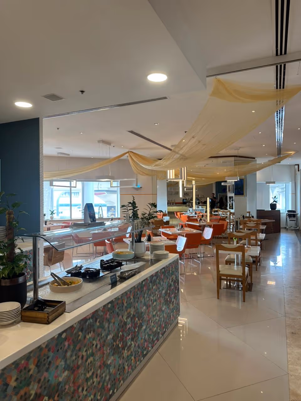 Modern restaurant interior with a buffet counter in the foreground, wooden chairs, and orange cushioned seats under ceiling drapes.