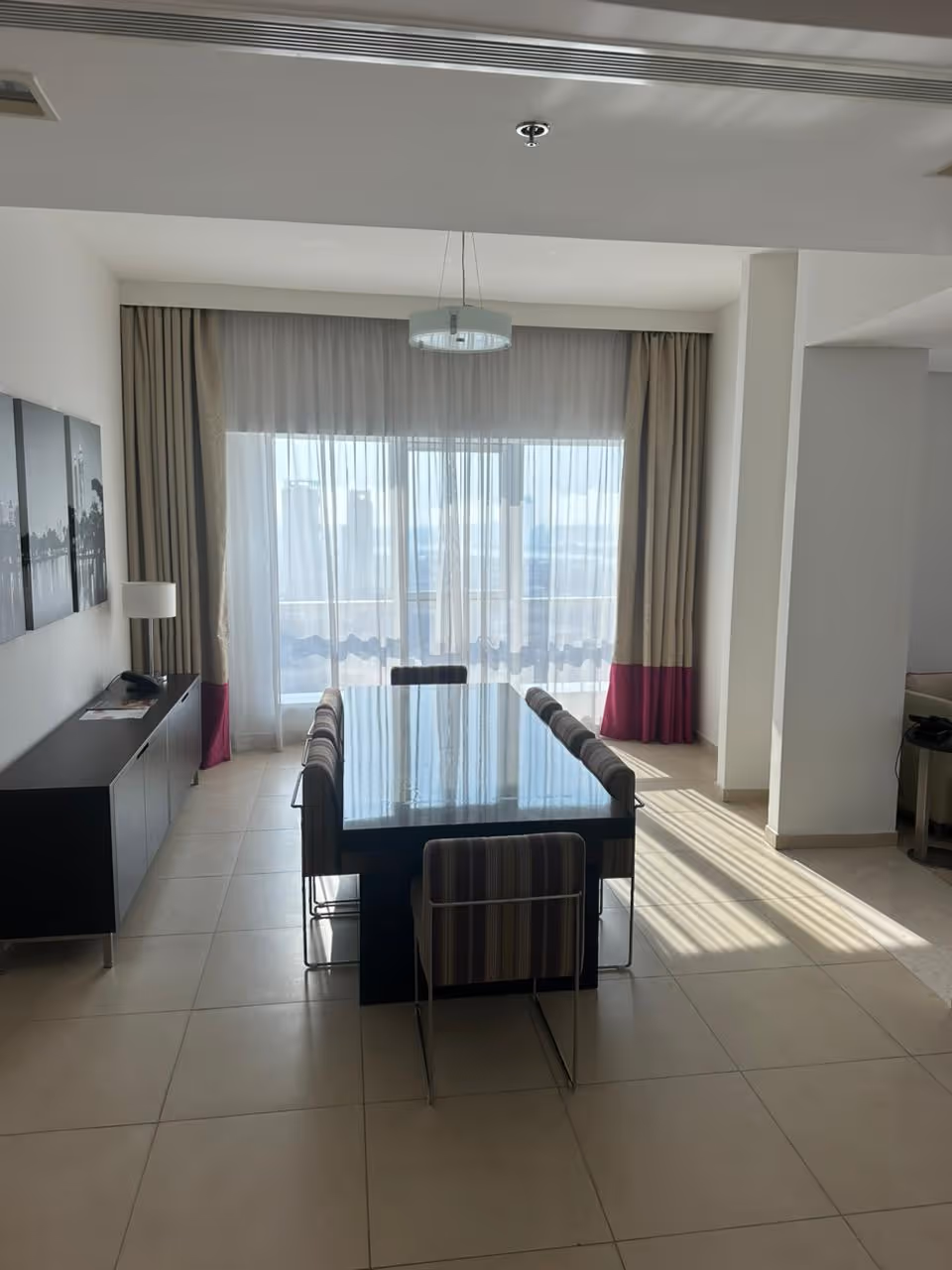 Living room with beige and white sofas facing a wall-mounted TV, a glass dining table with four transparent chairs, wooden floor, and decorative chandeliers on the ceiling.
