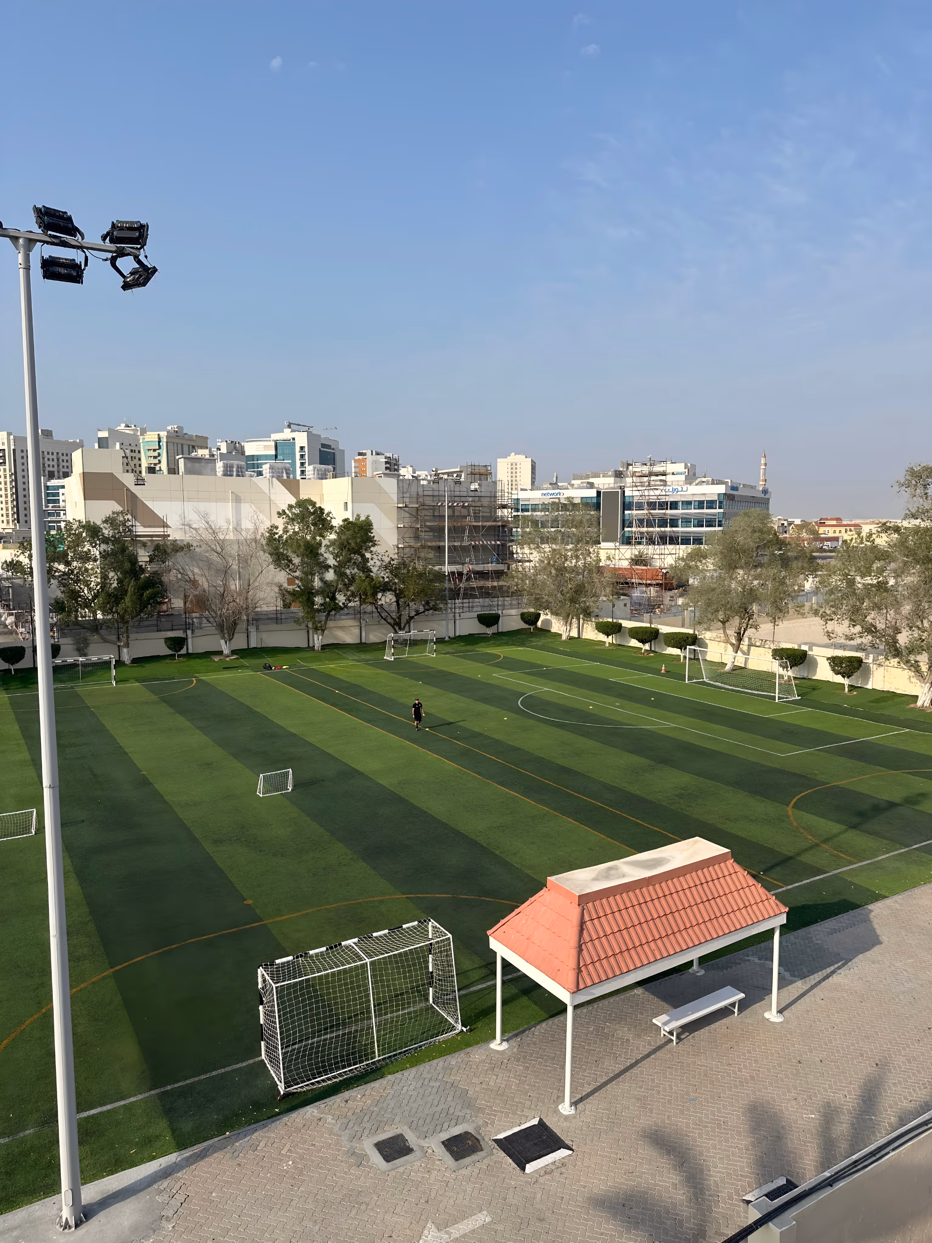 Outdoor sports field with green artificial turf, three soccer goals, a tall light pole, and surrounding buildings under a clear sky.