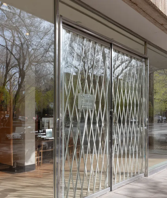 Glass entrance door with a metal accordion security gate, reflecting bare tree branches and surrounding street.