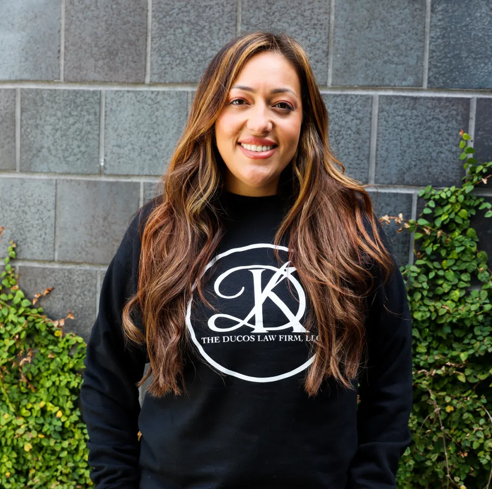 Smiling woman with long brown hair wearing a black sweatshirt with 'The Ducos Law Firm, LLC' logo, standing in front of a gray stone wall with green vines.