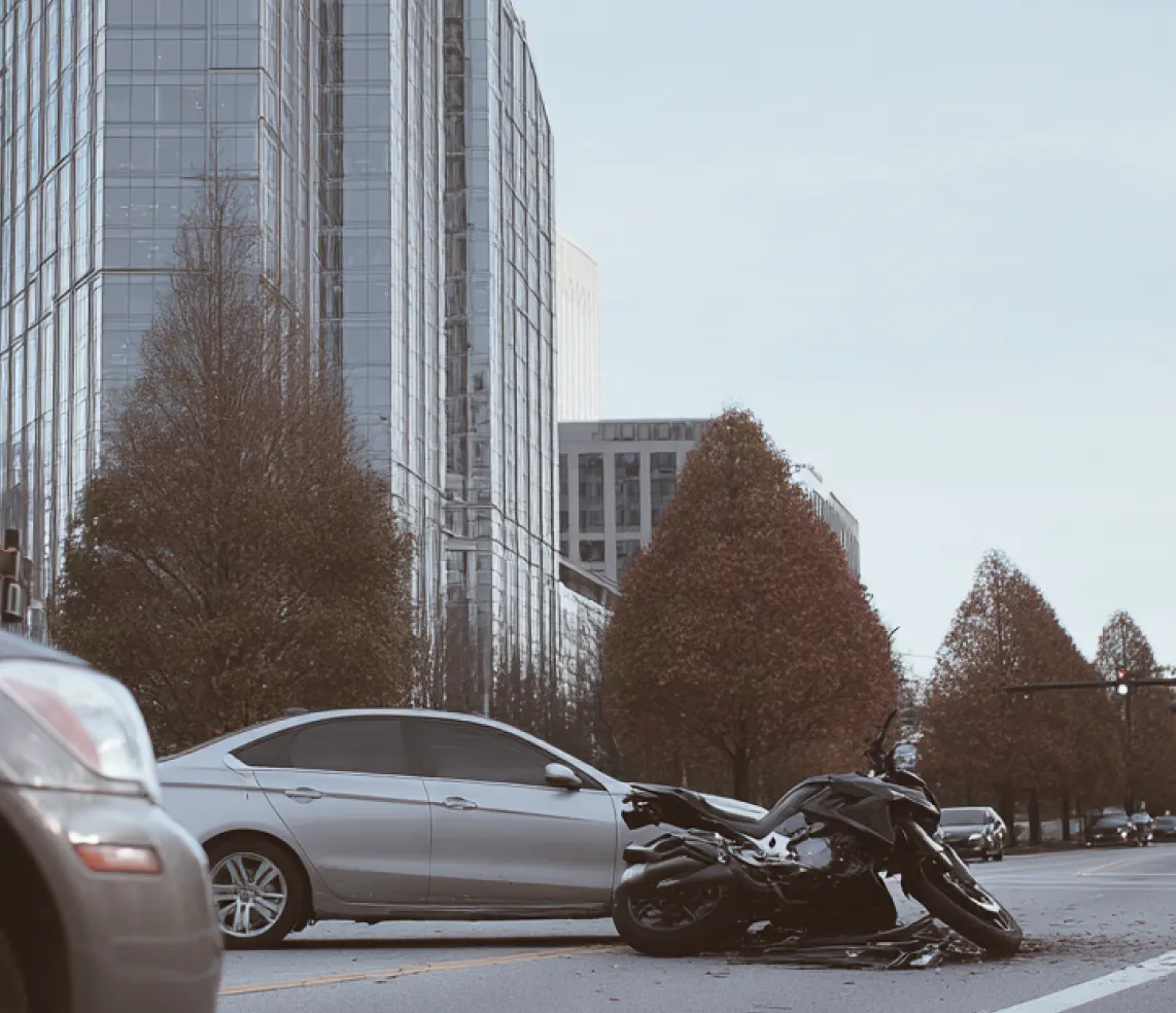 Black motorcycle lying on its side on a city street after a collision with a silver sedan, with glass debris scattered nearby and tall buildings and trees in the background.