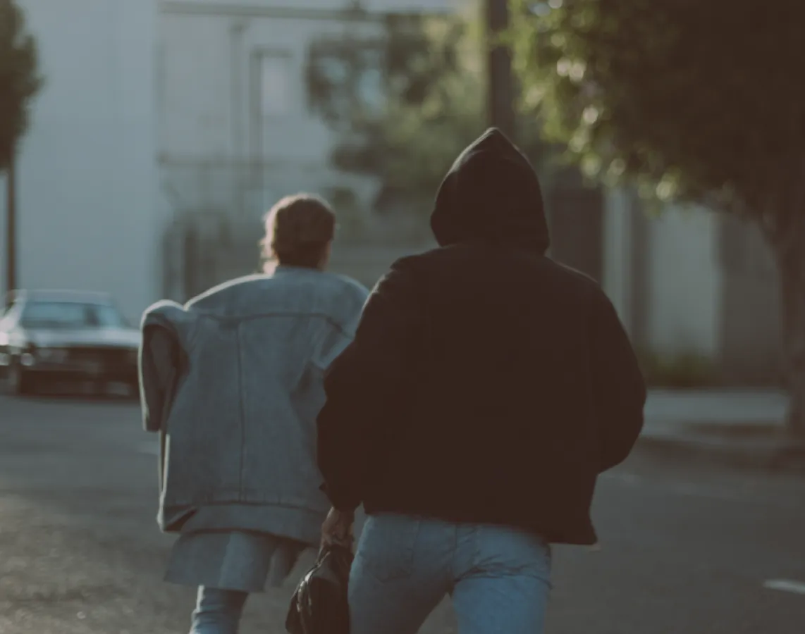 Two people walking away on a street, one wearing a denim jacket and the other a black hooded jacket.