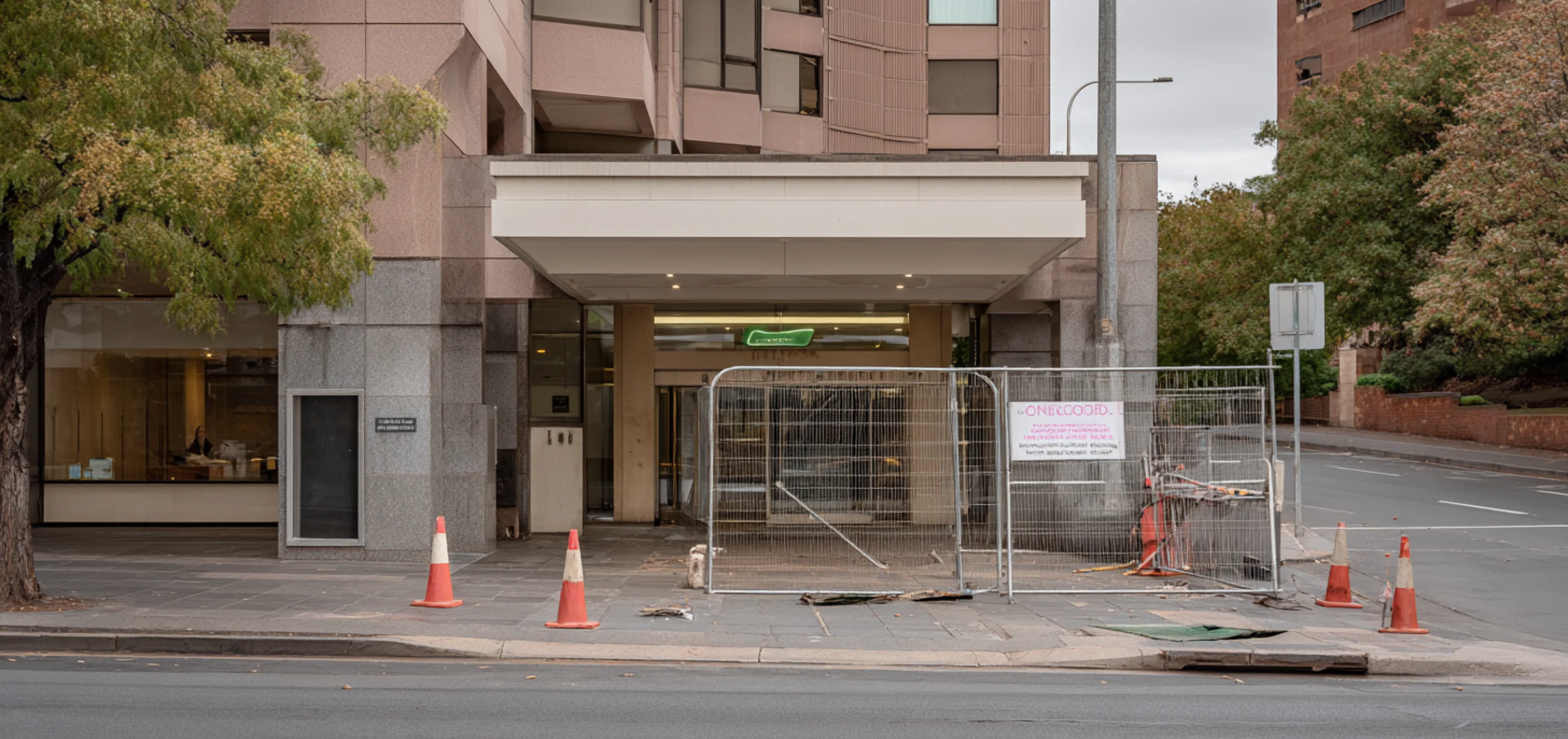 Entrance of a building blocked off with metal fencing and traffic cones indicating sidewalk construction work.