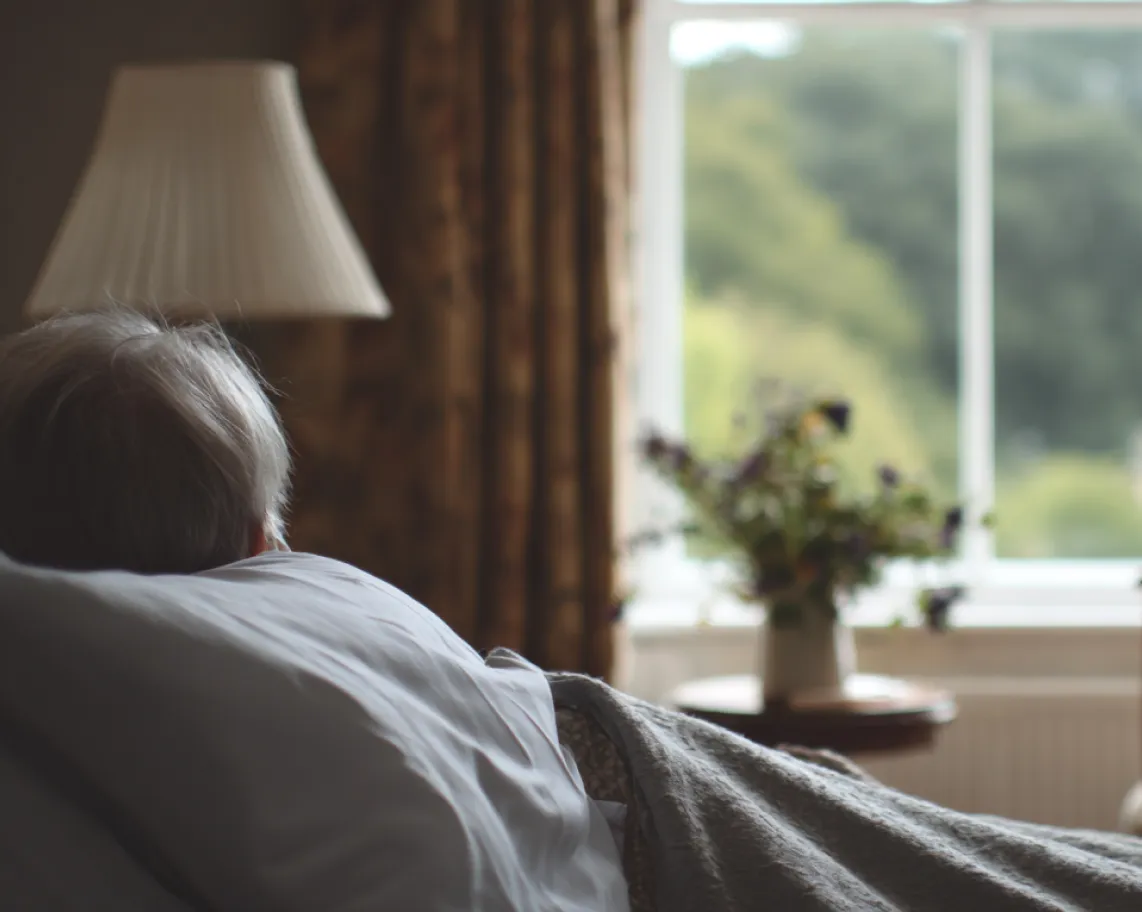 Elderly person lying in bed facing away, with a lit lamp and a vase of flowers by a window in the background.