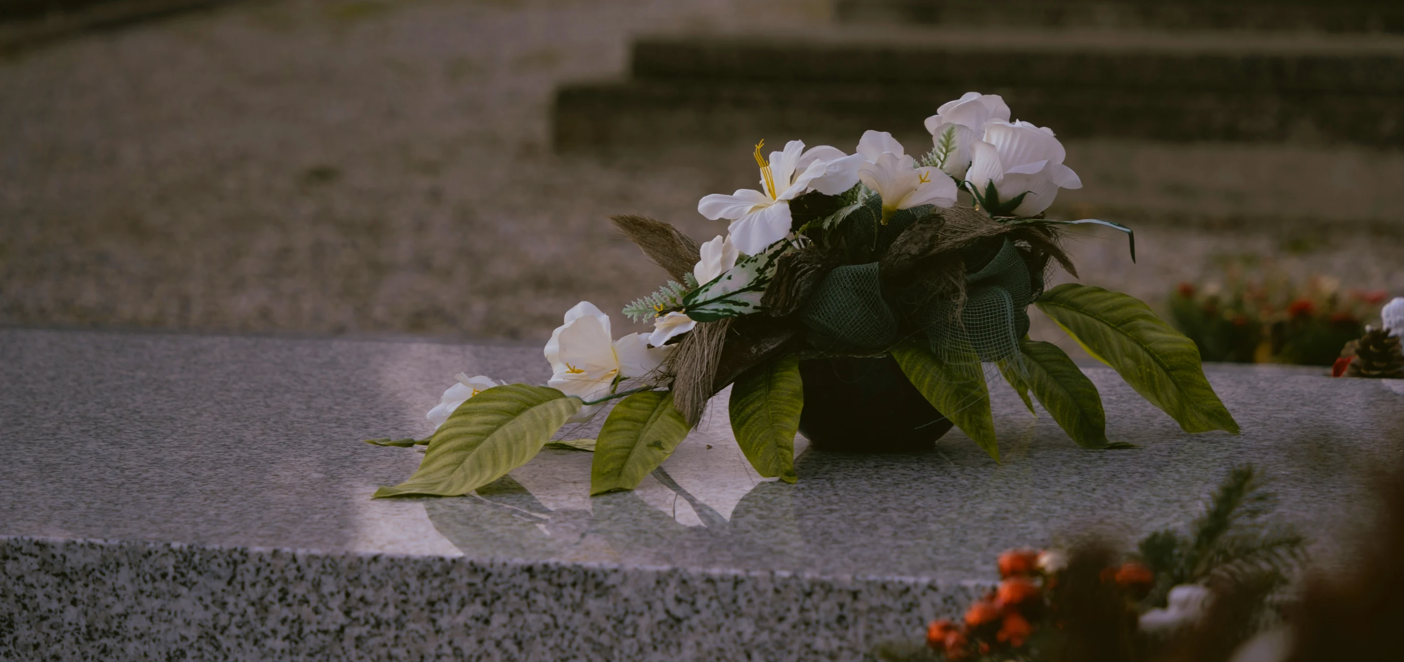 Artificial white flowers with green leaves arranged on a polished granite gravestone.