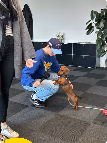 A man in a blue jacket and baseball cap kneels on tile flooring holding a microphone, while a small dachshund rests its front paws on his knees.