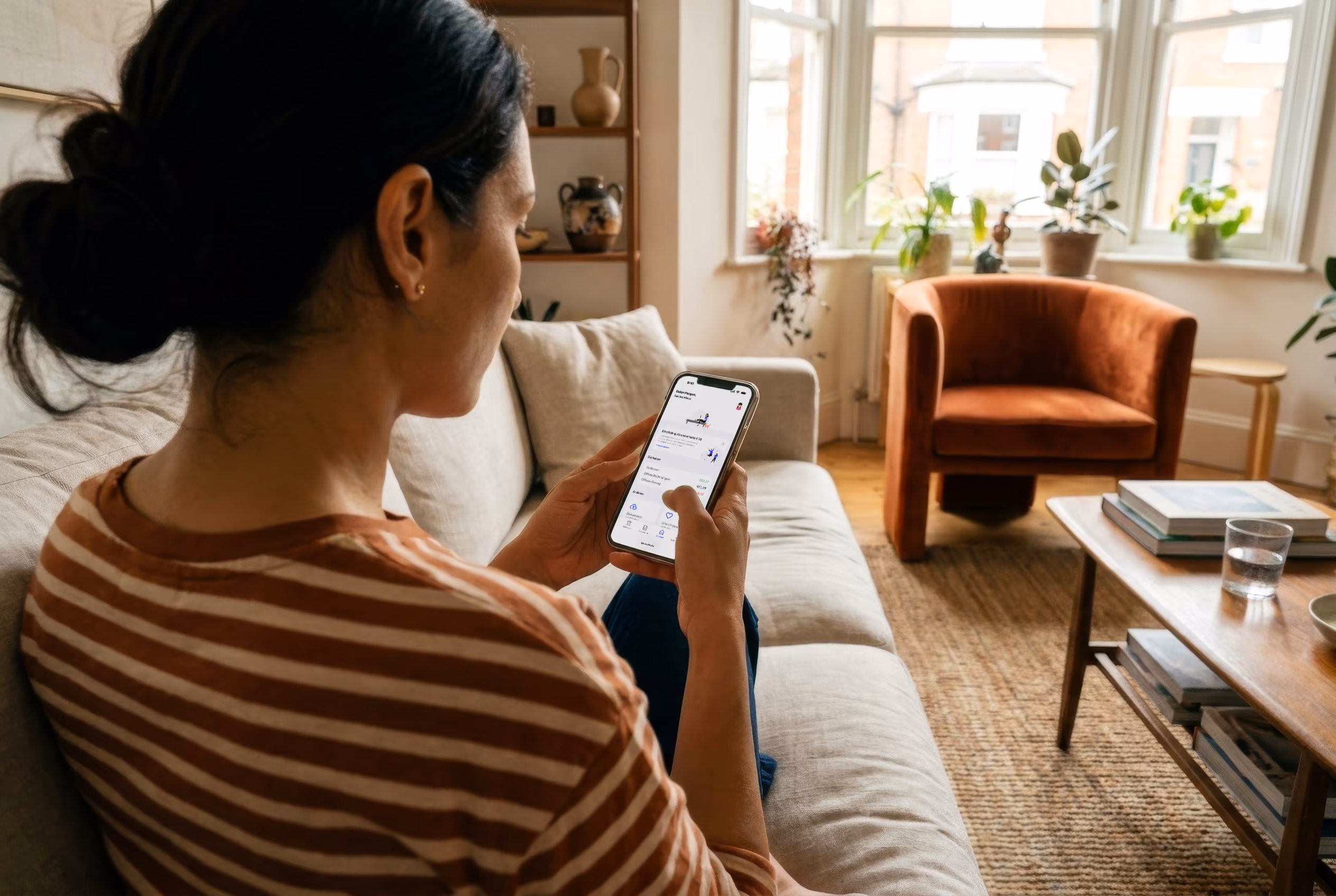 Woman sitting on a sofa using her smartphone in a bright living room with plants and an armchair.