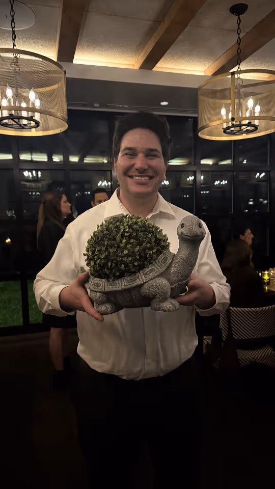 Smiling man in a white shirt holding a decorative turtle planter with greenery inside in a warmly lit room.