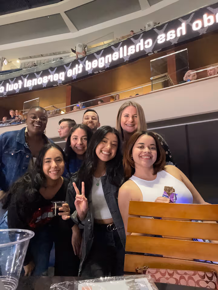 Group of seven diverse friends smiling and posing for a photo at an indoor event with balcony seating above.