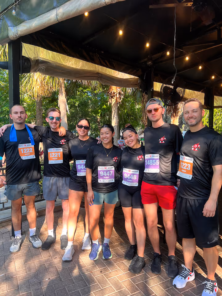 Group of seven runners wearing black shirts and race bibs standing closely together outdoors under a canopy.
