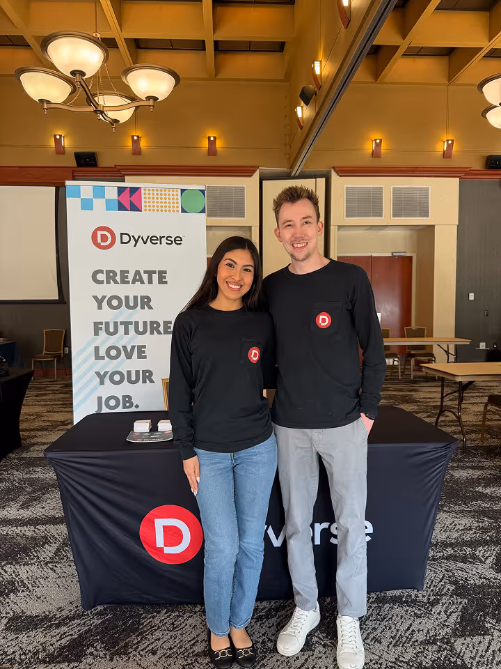 A smiling man and woman standing side by side in front of a Dyverse branded table and banner with the text 'Create your future. Love your job.'