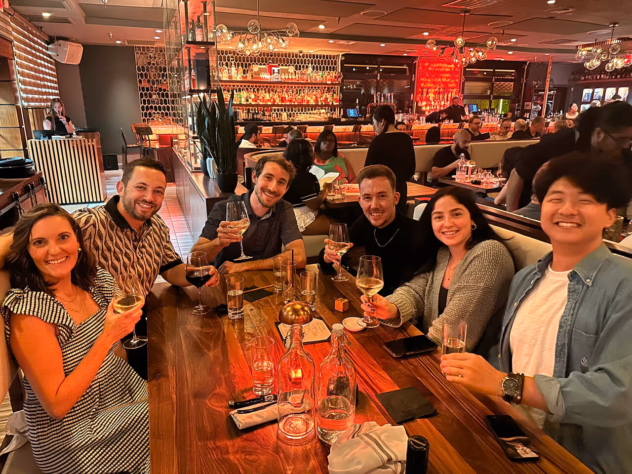 Six friends sitting around a wooden table in a warmly lit restaurant, smiling and holding glasses of wine or drinks.