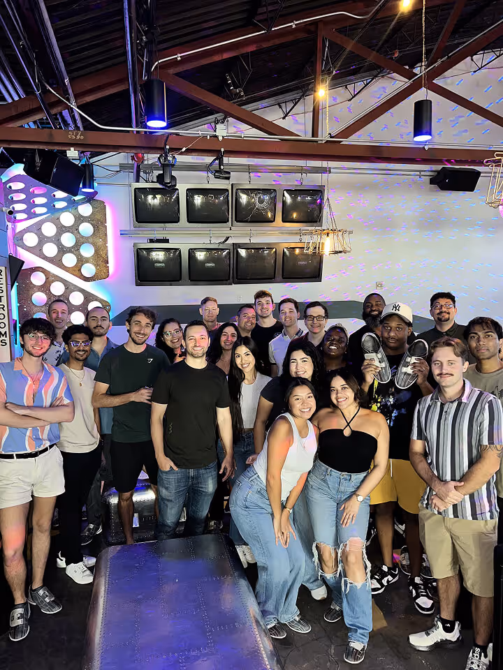 A diverse group of young adults smiling and posing together indoors, with modern lighting and decor in the background.