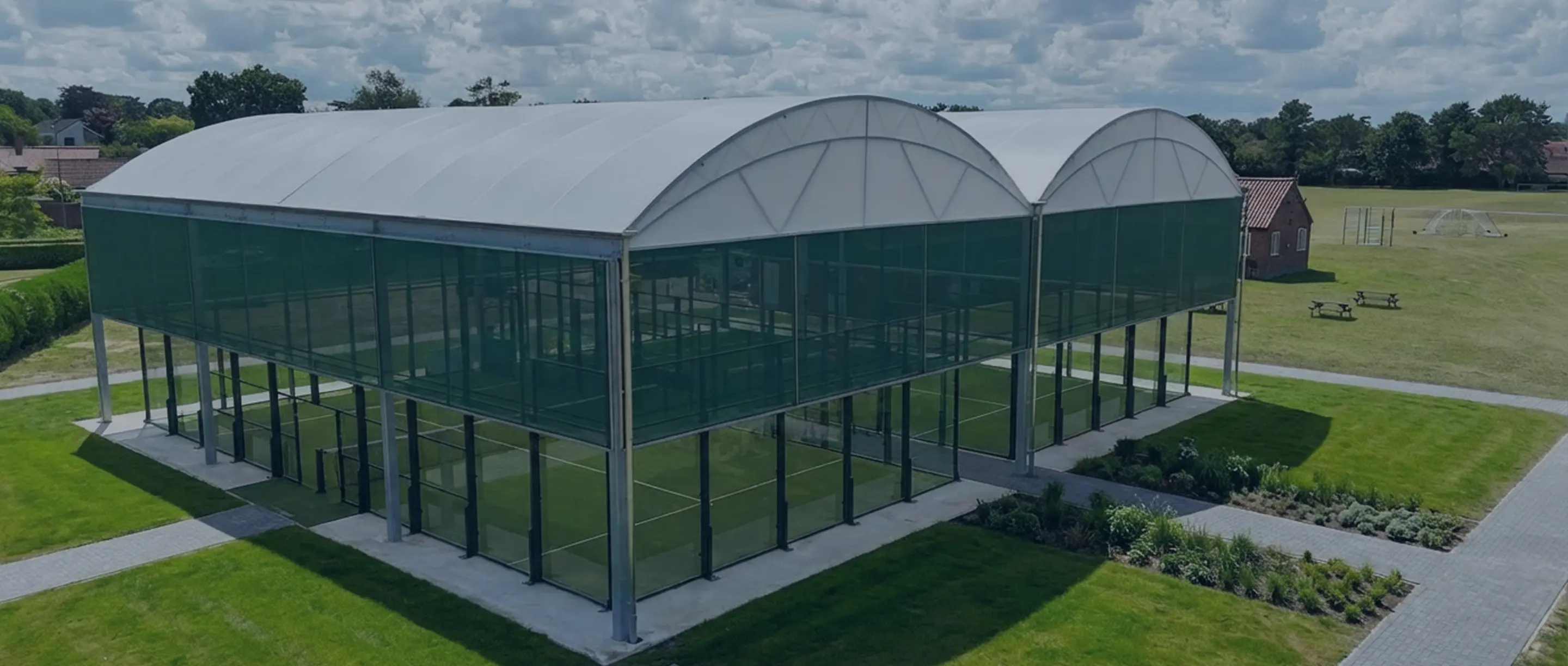 Modern outdoor sports courts enclosed by transparent green mesh and white arched roofs in a grassy park area with nearby walking paths and picnic tables.