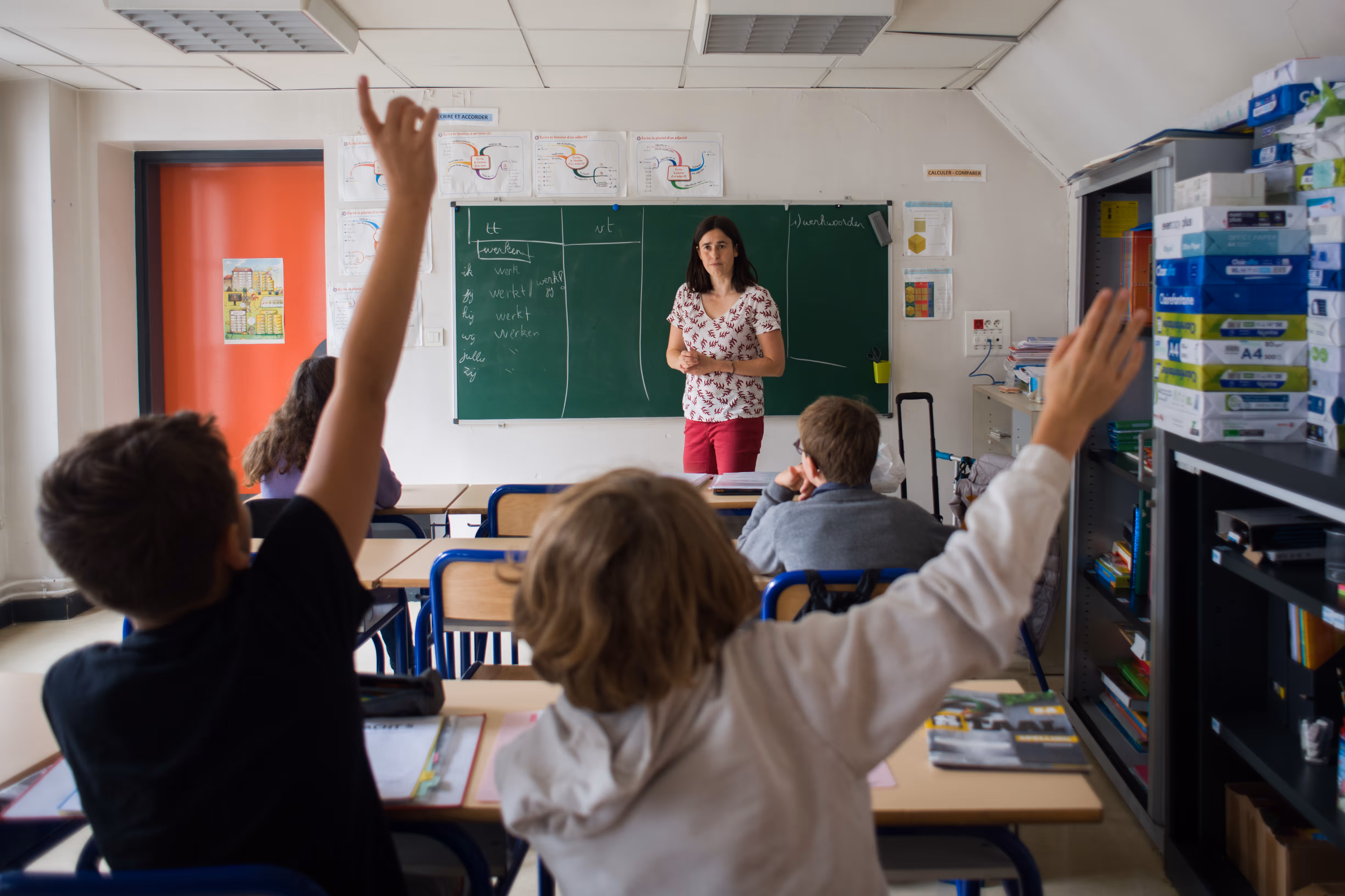 Basisschoollerares geeft les aan kinderen, twee leerlingen steken handen op in klaslokaal.