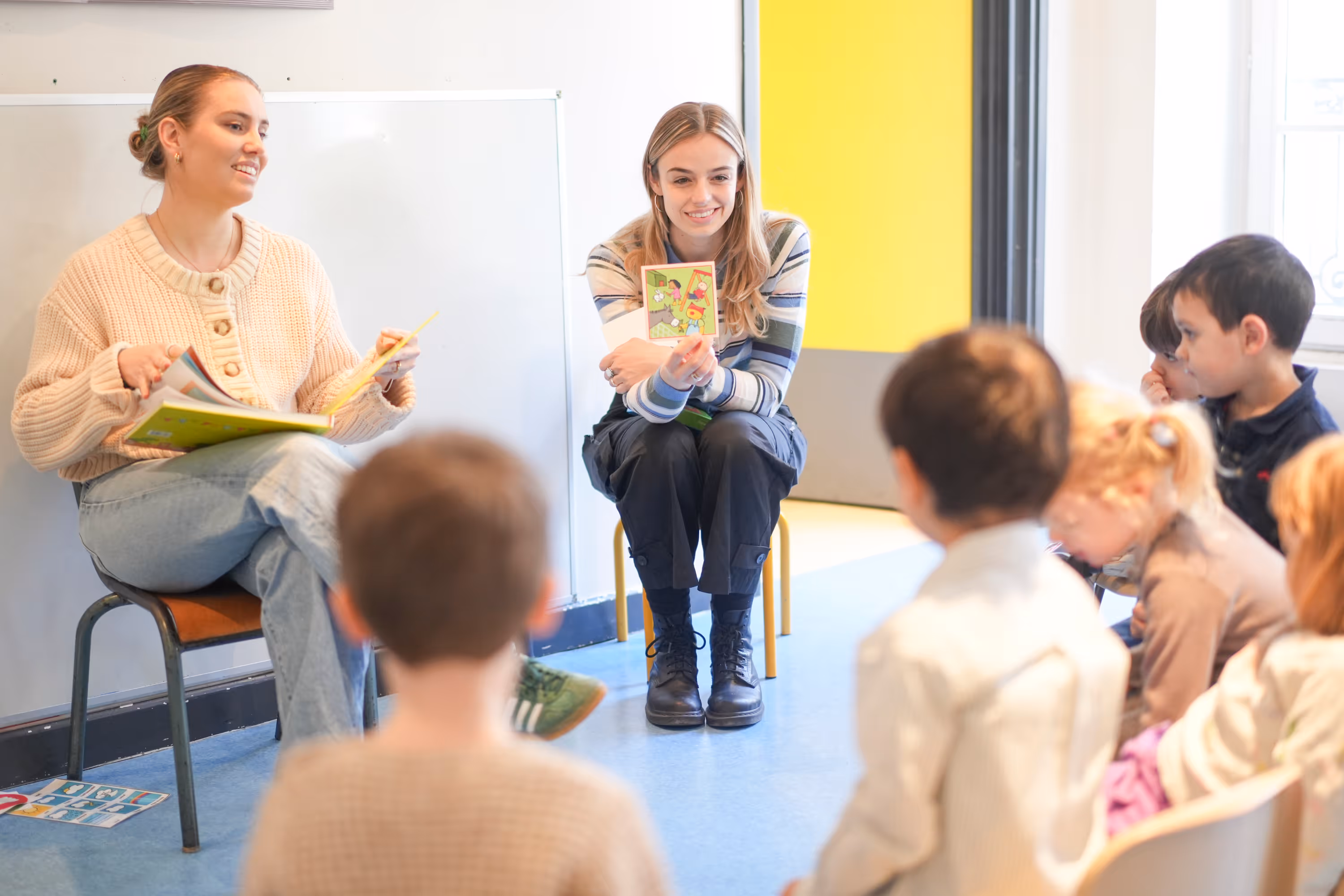 Twee vrouwen lezen voor aan een groep jonge kinderen die aandachtig luisteren in een klaslokaal.