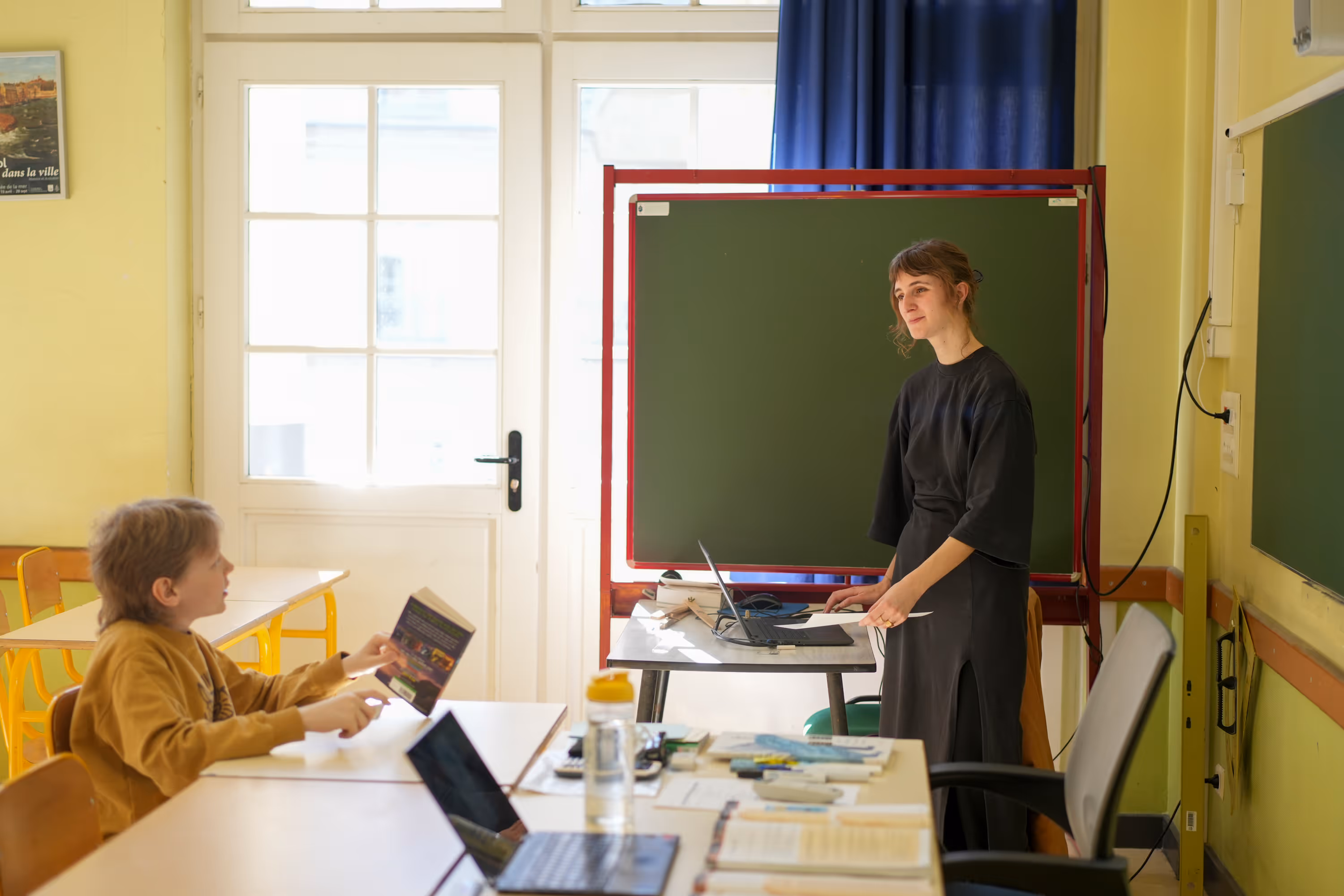 Lerares in een klaslokaal spreekt met een leerling die een boek vasthoudt.