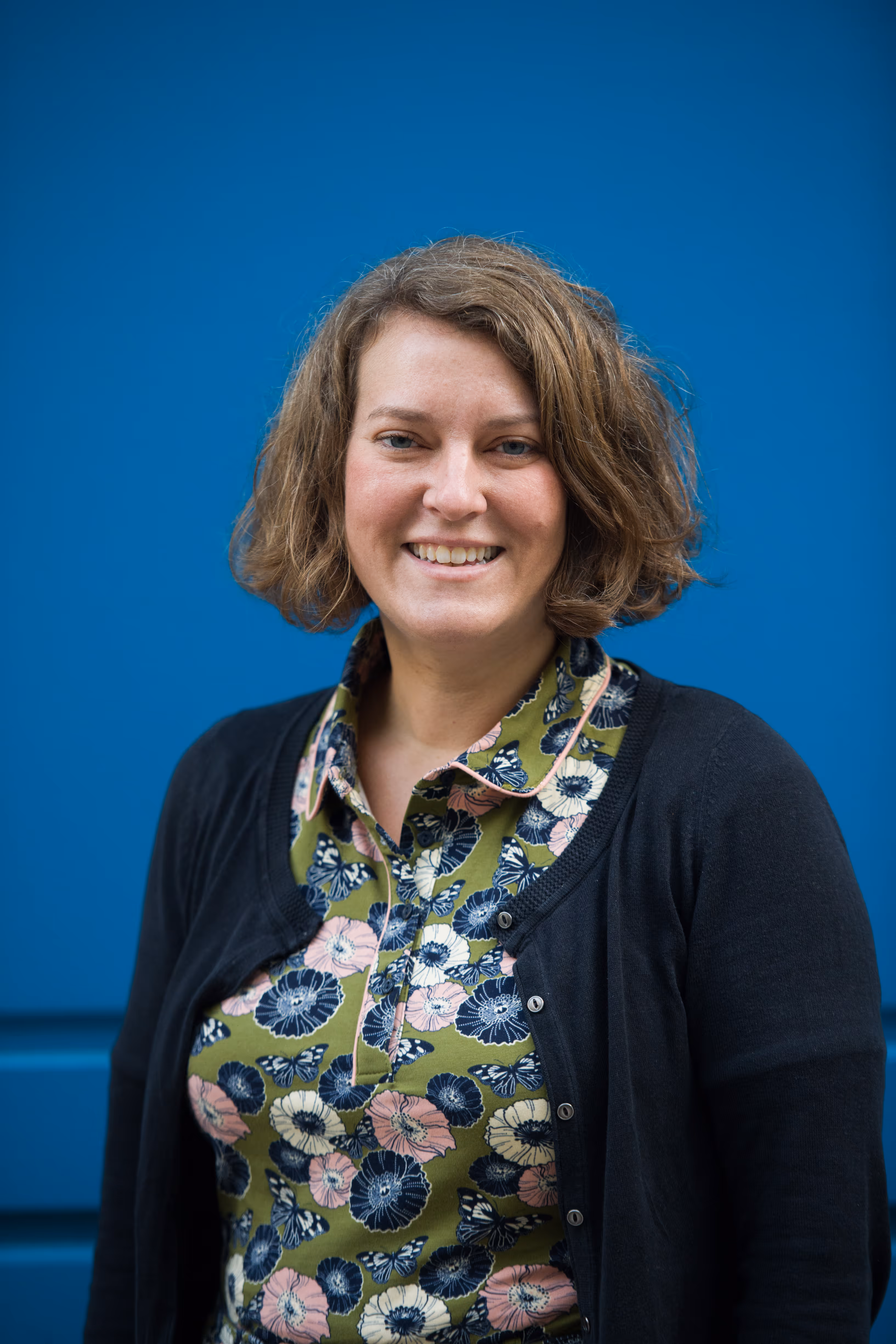 Vrouw met kort, golvend haar, glimlachend met een bloemenblouse en zwarte cardigan voor een blauwe muur.