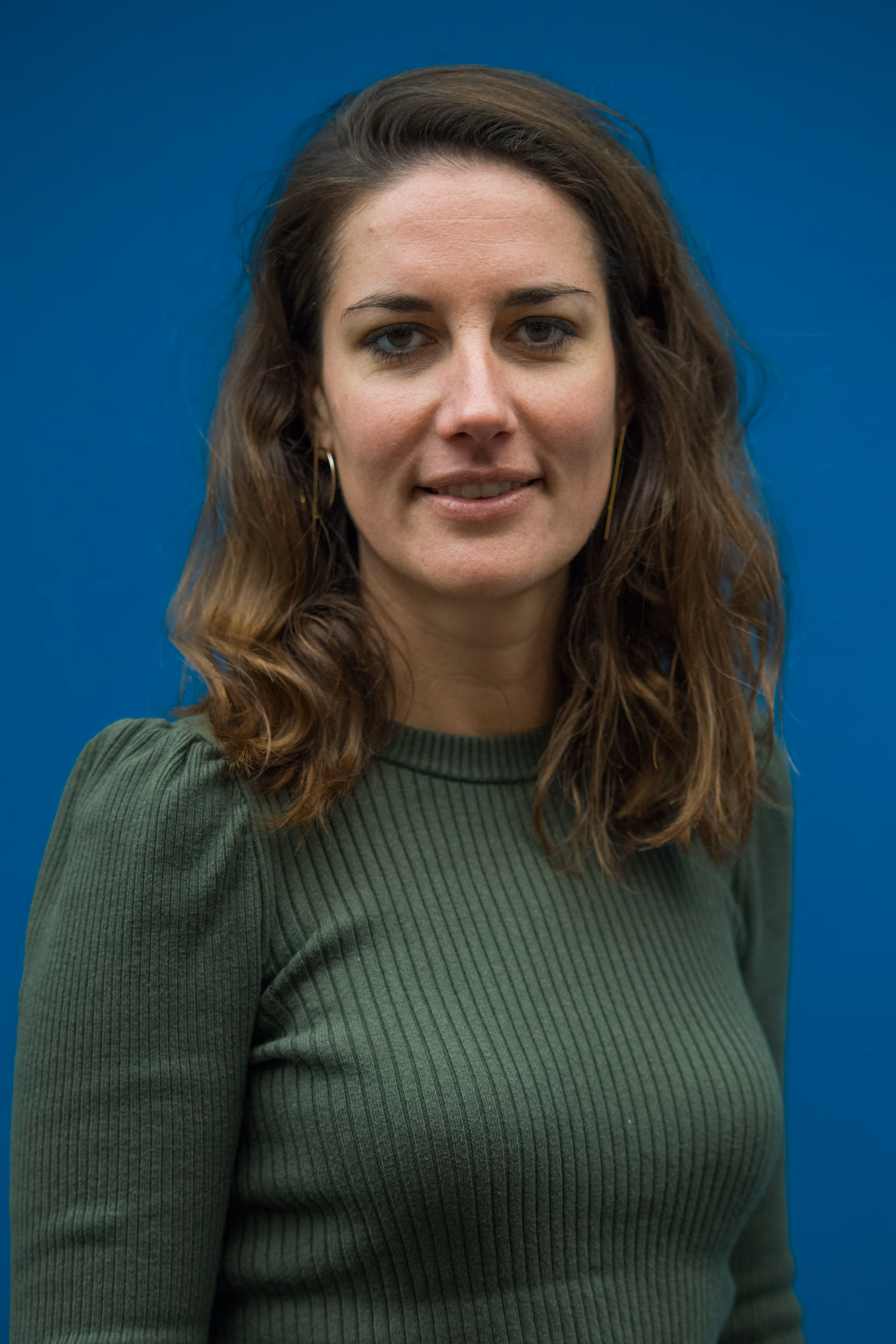 Vrouw met golvend bruin haar draagt een groene geribbelde trui en gouden oorbellen, blauwe achtergrond.