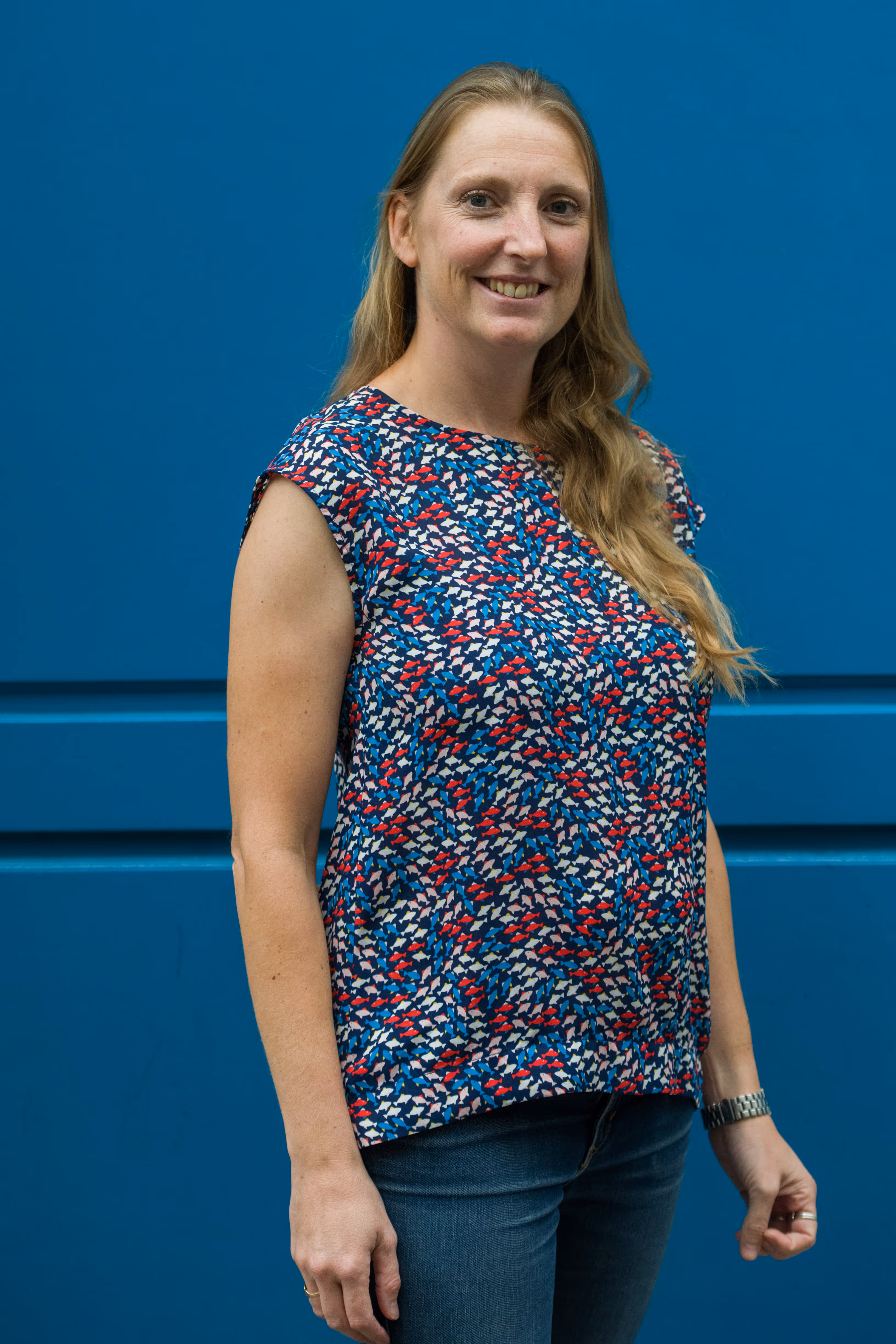 Vrouw met lang blond haar en een kleurrijk blouseje staat voor een blauwe muur en lacht.