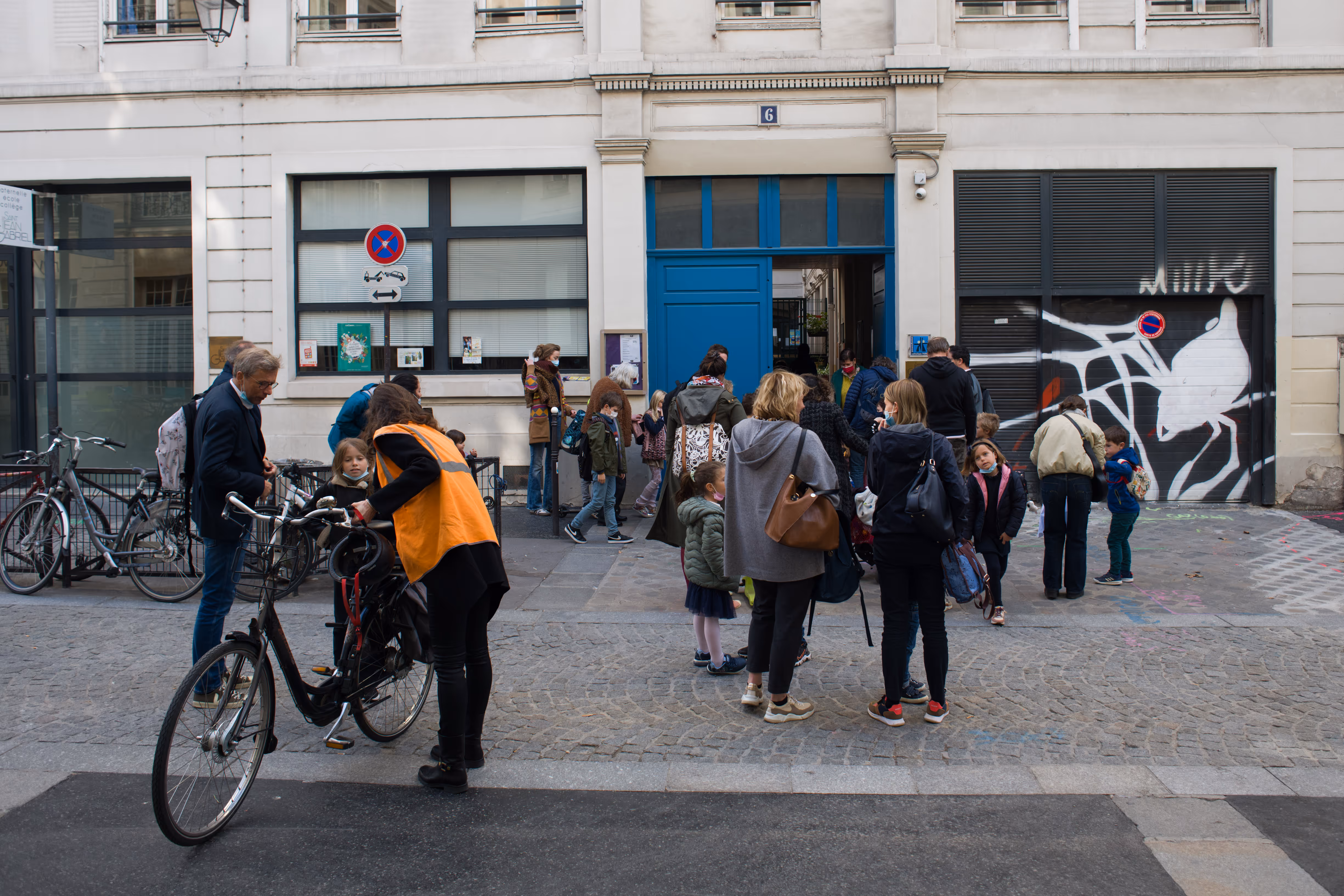 Volwassenen en kinderen verzamelen zich buiten een schoolgebouw met een blauwe deur in de straat.