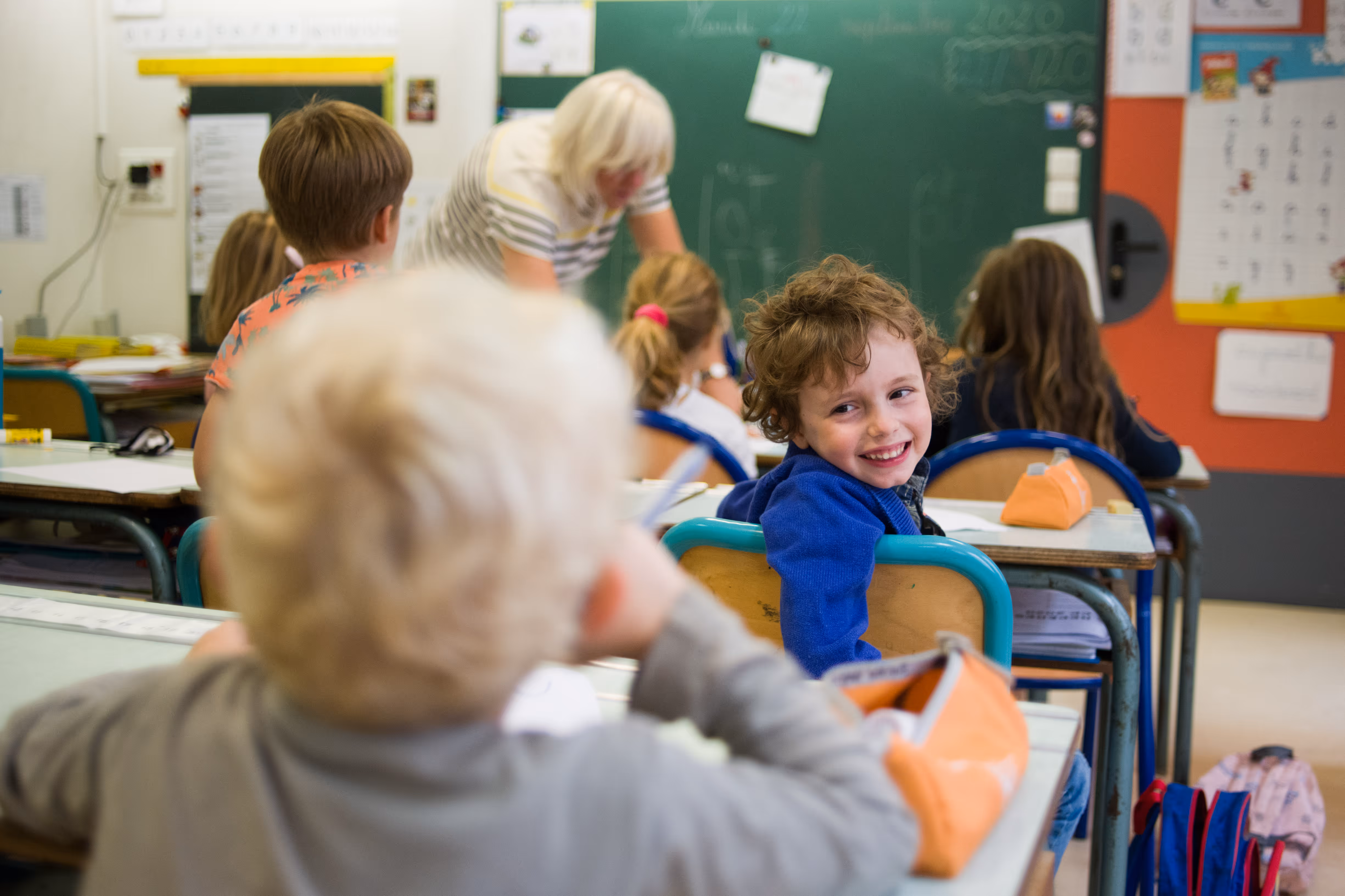 Lachend kind in blauw jasje kijkt naar klasgenoot in een basisschoolklaslokaal.