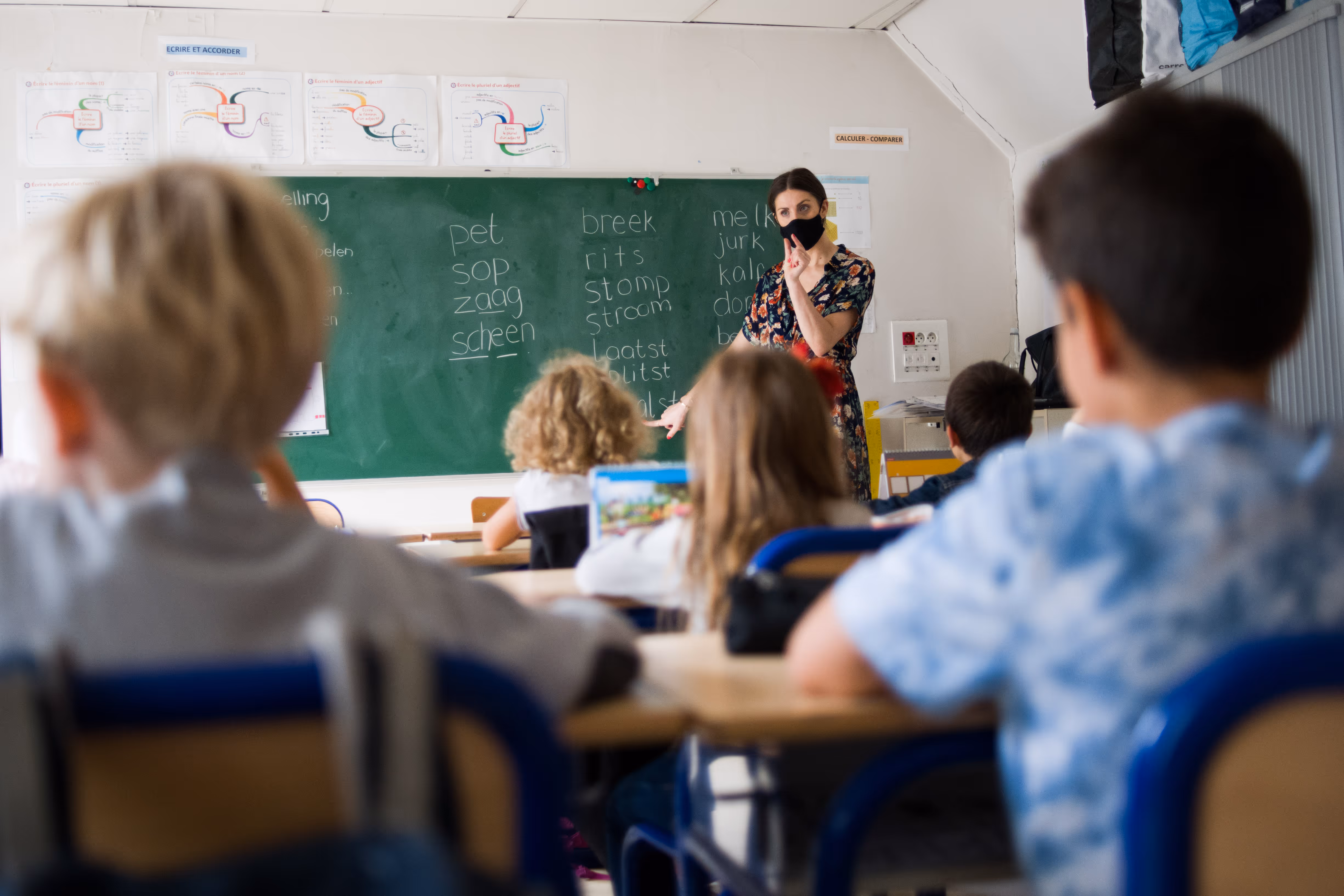 Juf met mondkapje geeft les aan jonge leerlingen bij bord met Nederlandse woorden.