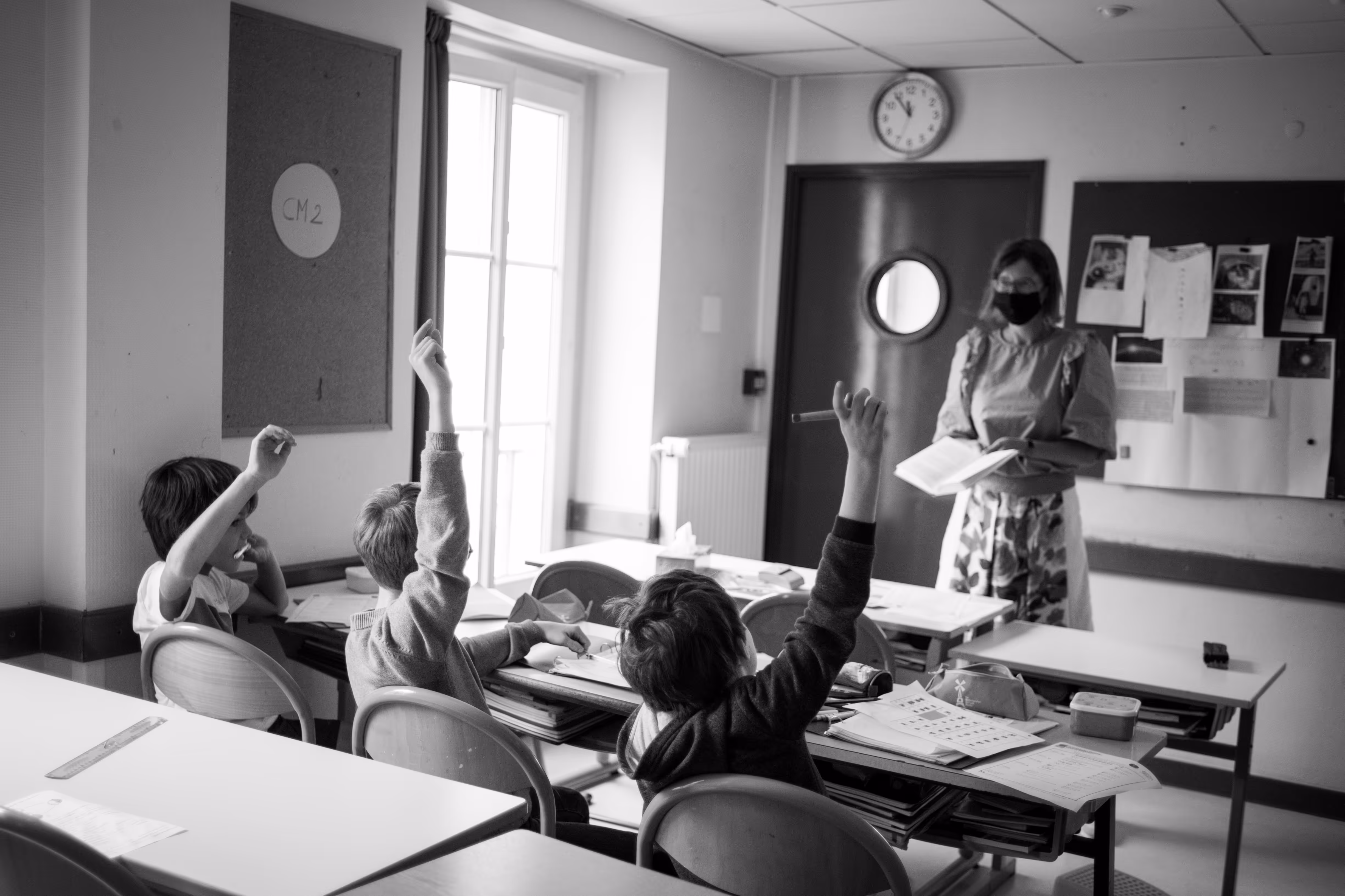 Drie kinderen in een klaslokaal steken hun hand op terwijl de lerares leest met mondkapje.