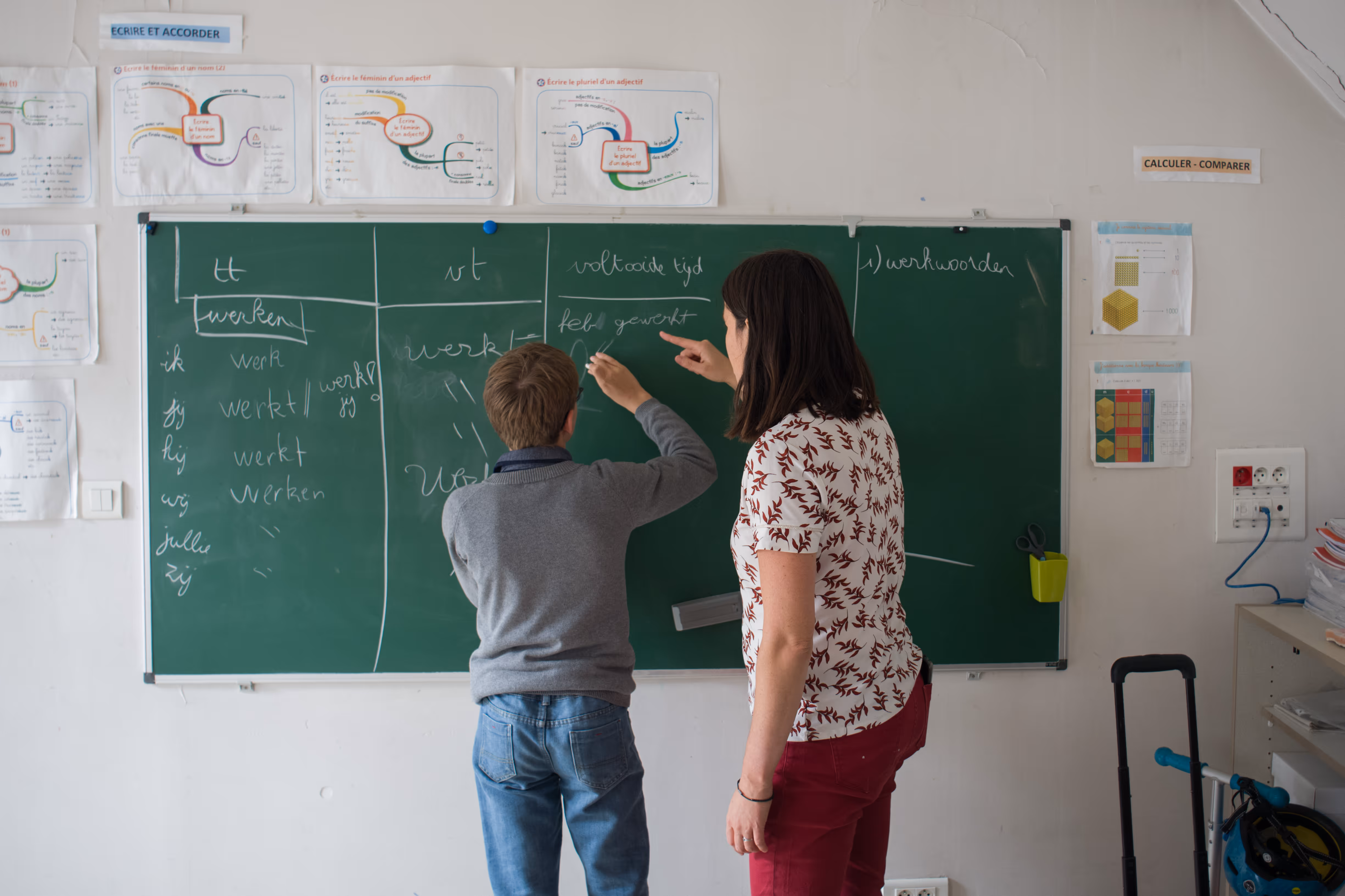 Juf helpt leerling schrijven op schoolbord met werkwoordvervoeging in klaslokaal.
